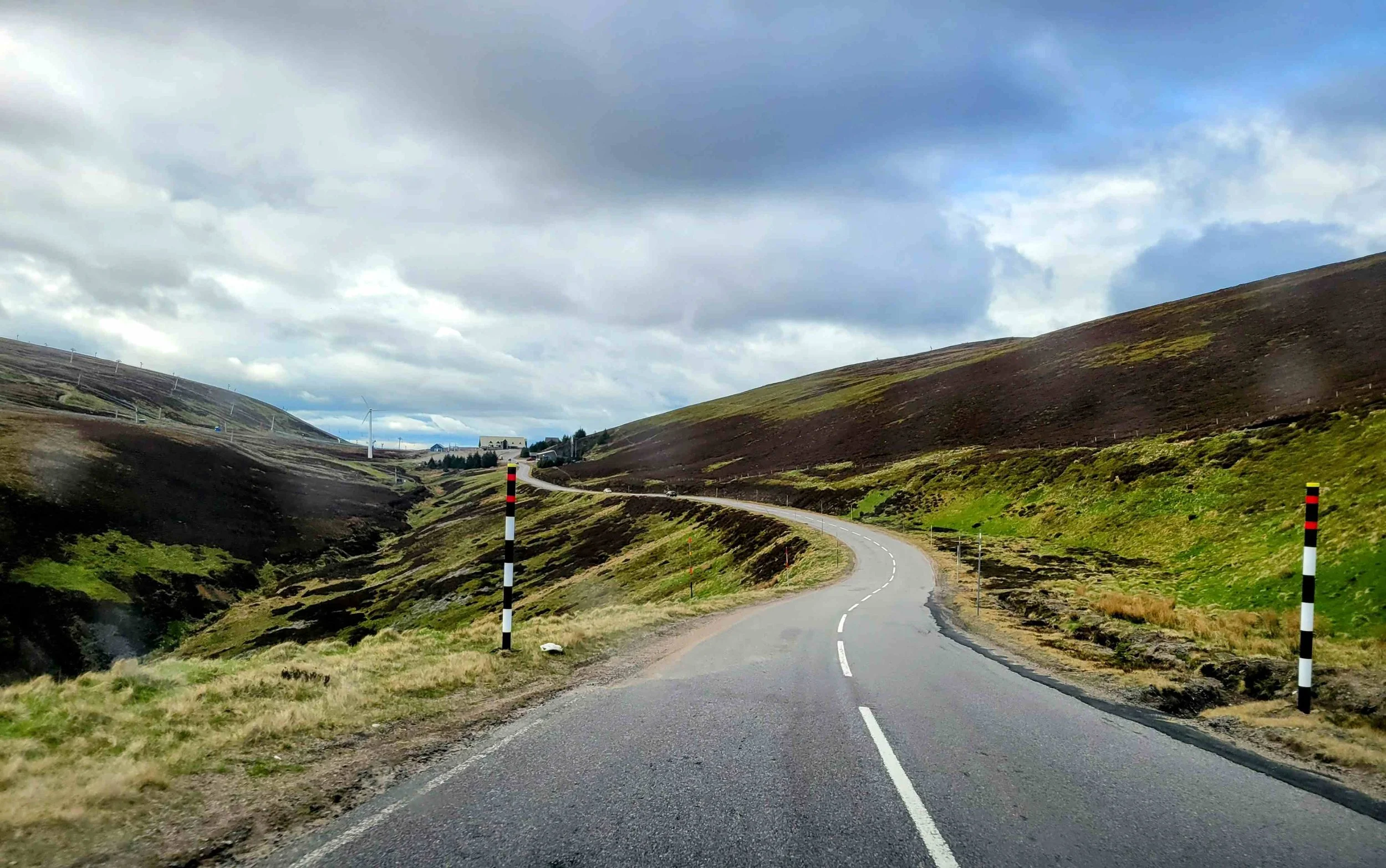A winding road through a hilly landscape with green and brown vegetation, under a cloudy sky.