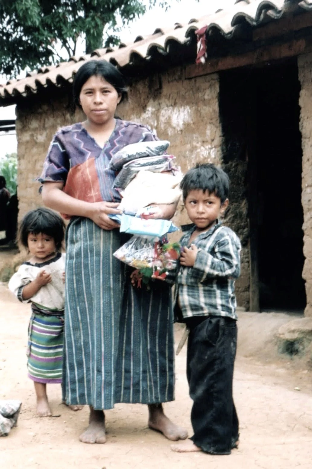 A woman holding several bags of supplies standing outside a rustic mud brick house with two children nearby, one boy and one girl, in a rural setting.