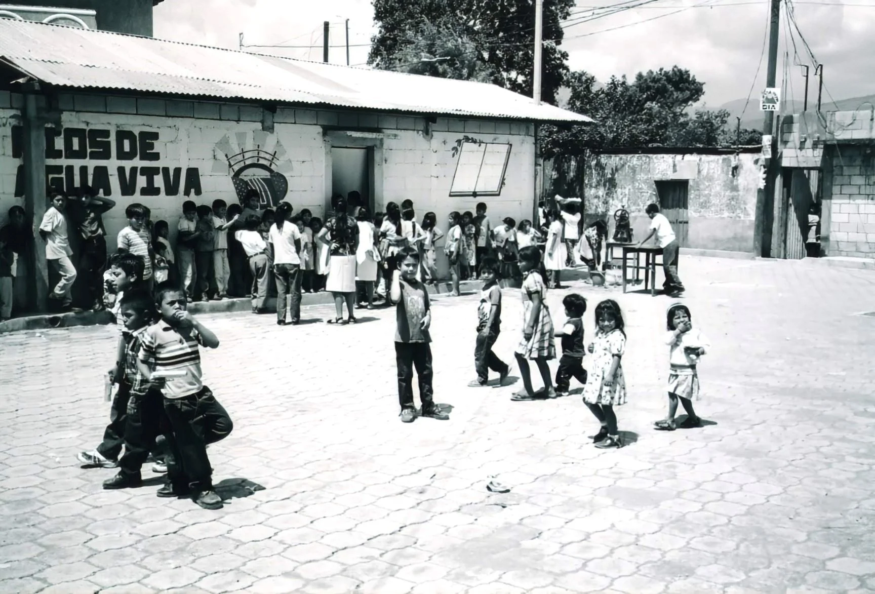 Children and students standing and walking outside a school building with a brick wall that has a sign in Spanish, in a rural area with cobblestone ground and utility poles.