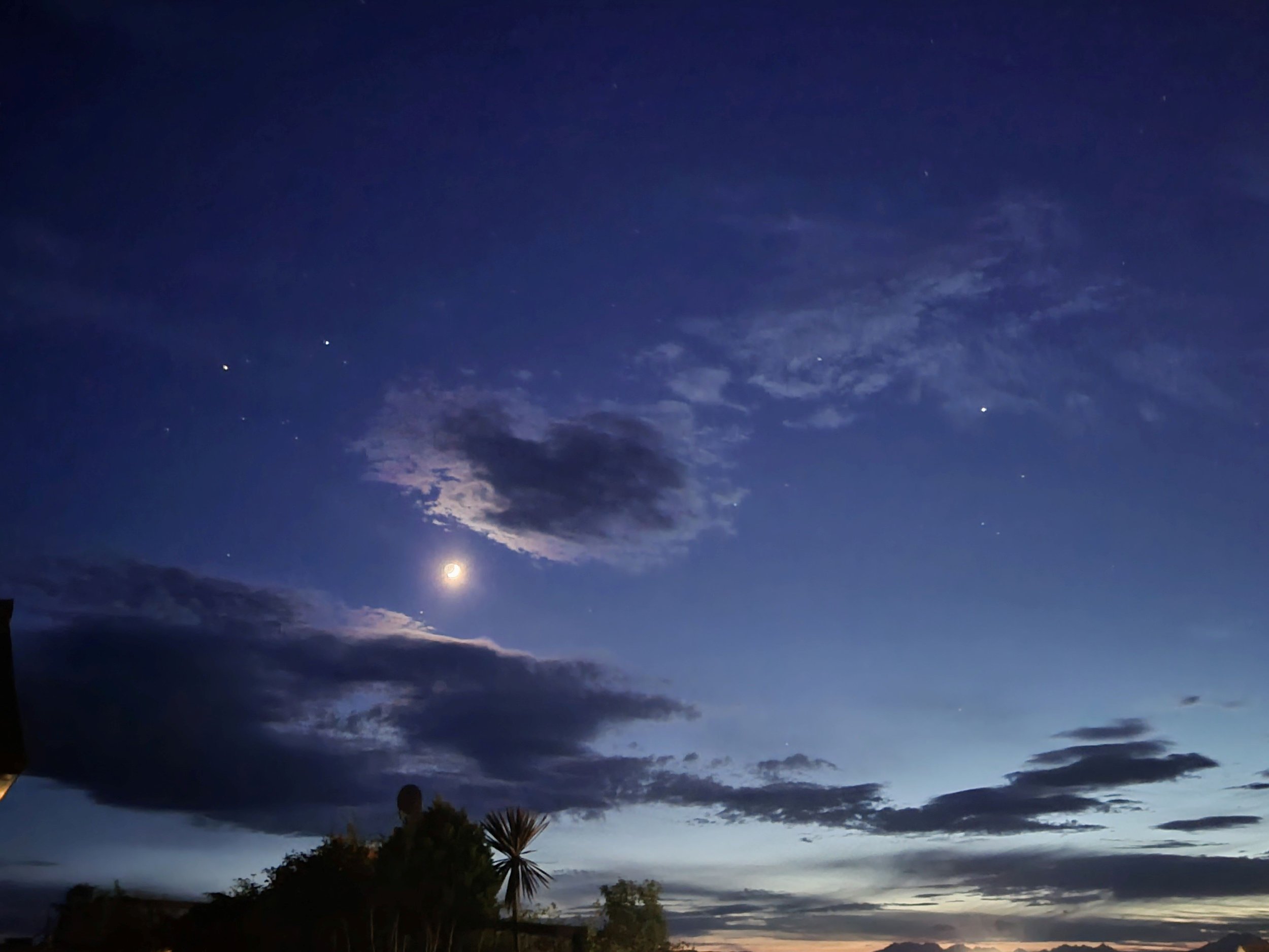Night sky with clouds and visible moon and stars above silhouetted trees. Midnight in Mallaig.