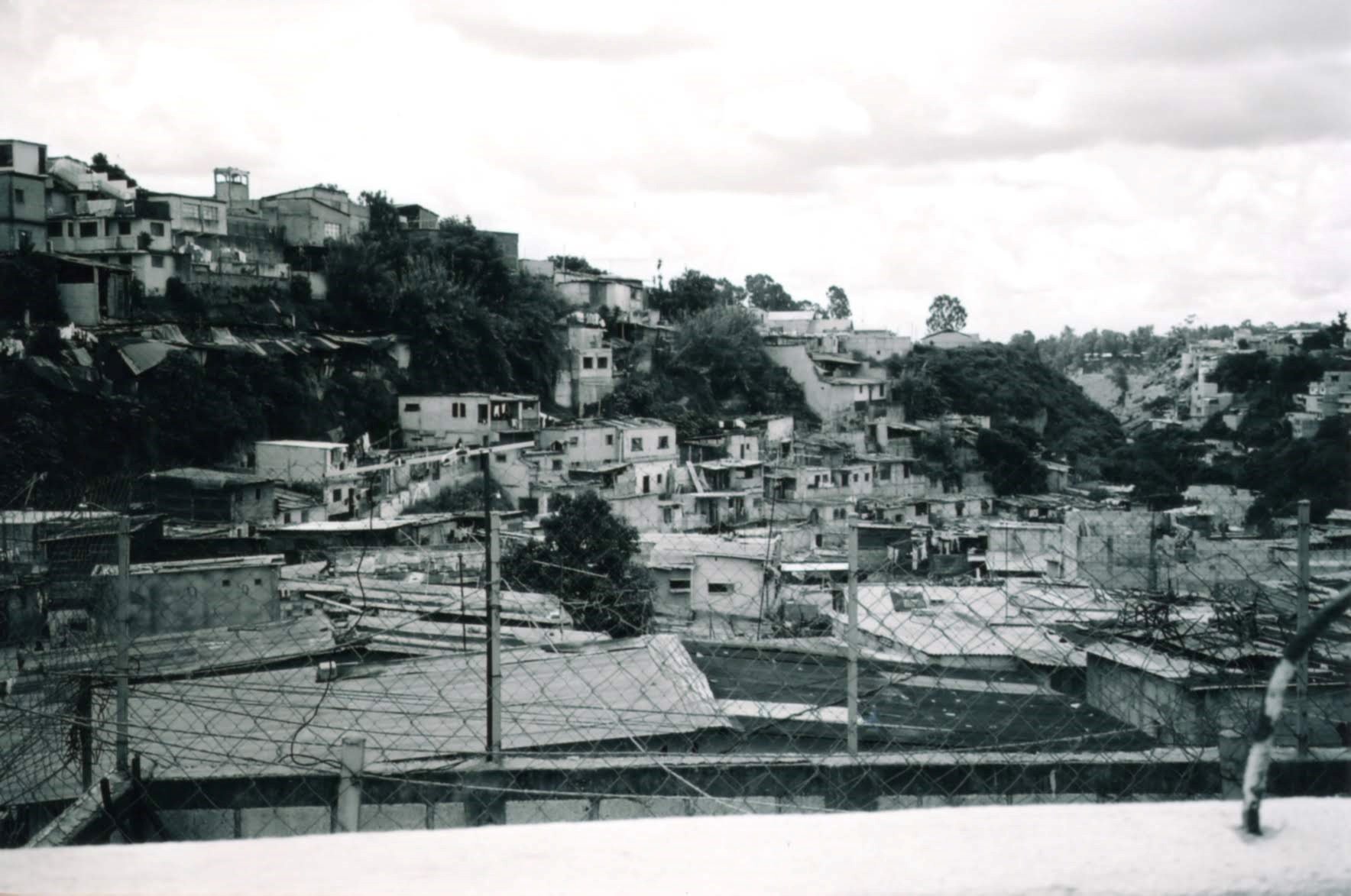 A hillside community with densely packed houses, some with flat roofs, surrounded by trees and a cloudy sky, viewed through a chain-link fence.