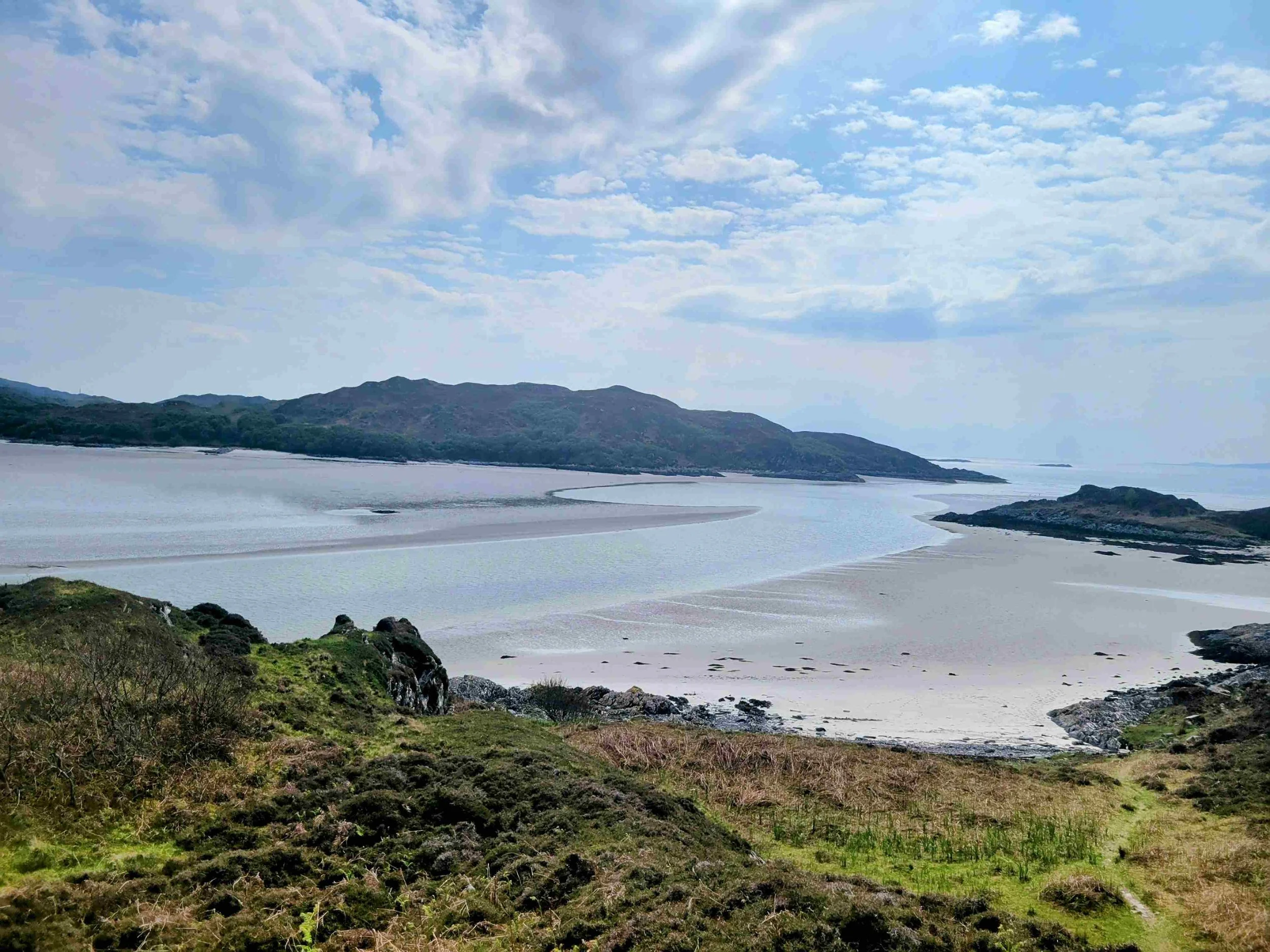 View of a landscape with a large water body, surrounding hills, and a partly cloudy sky.