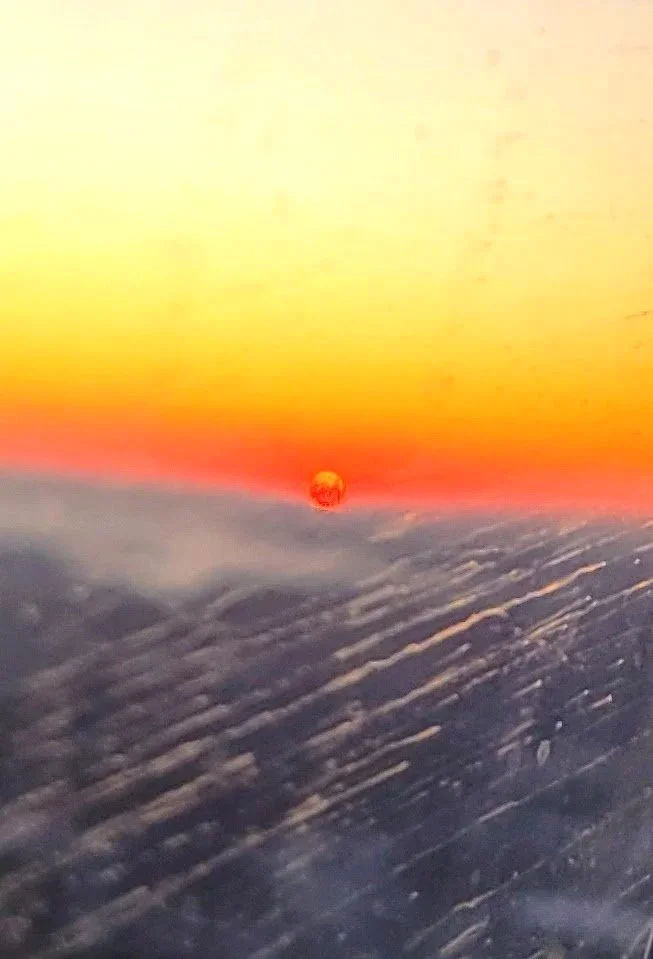 View from an airplane window showing a sunset or sunrise with the sun near the horizon, partially obscured by clouds and atmospheric haze.