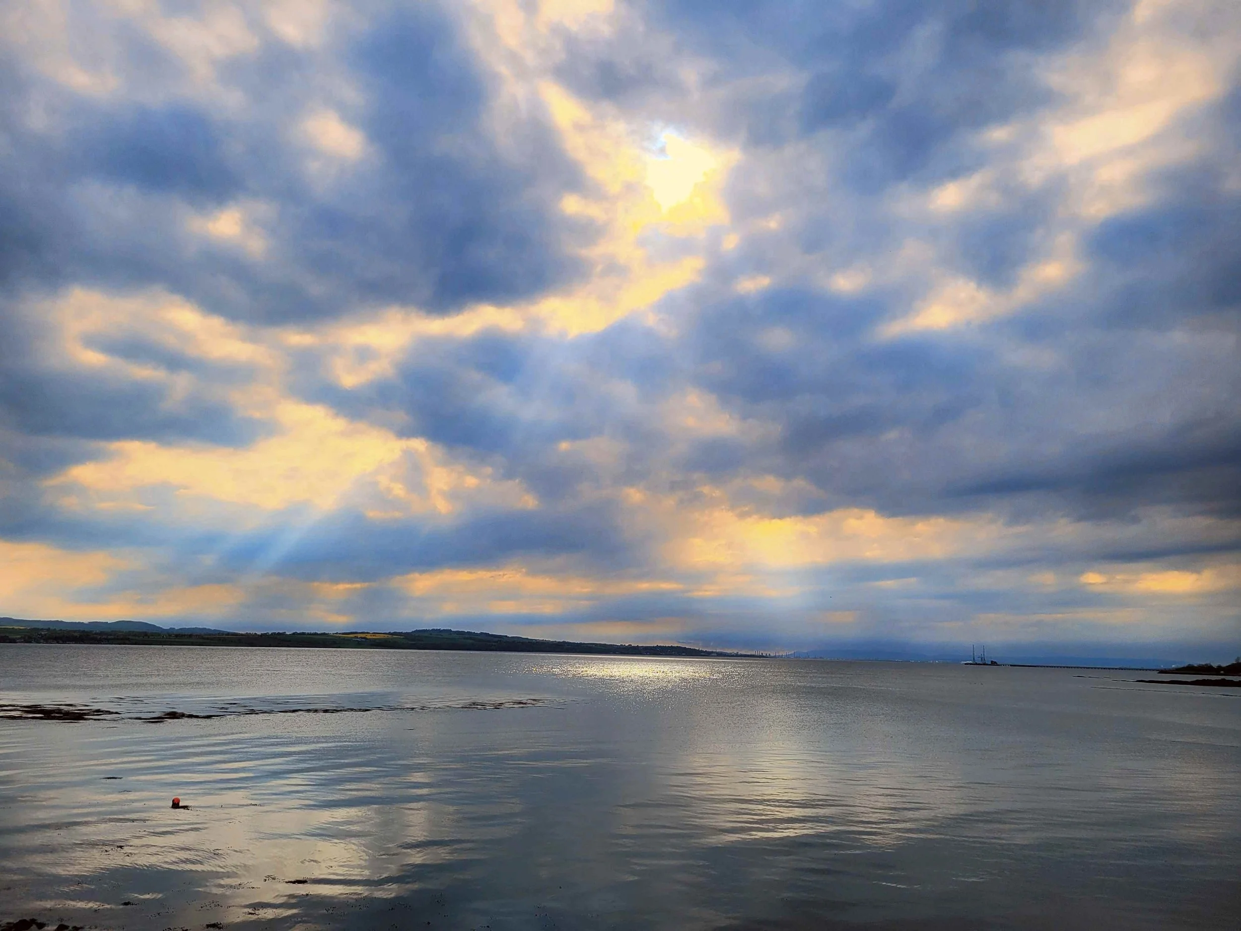 A large body of water reflects a cloudy sky at sunset, with a shoreline and a tiny buoy floating in the water.  Limekilns, Scotland
