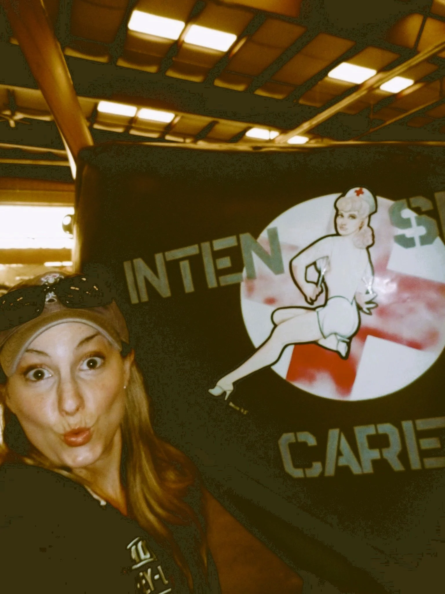 A woman with long red hair posing for a selfie, with a cartoon-style pin-up nurse on the tail of an airplane.