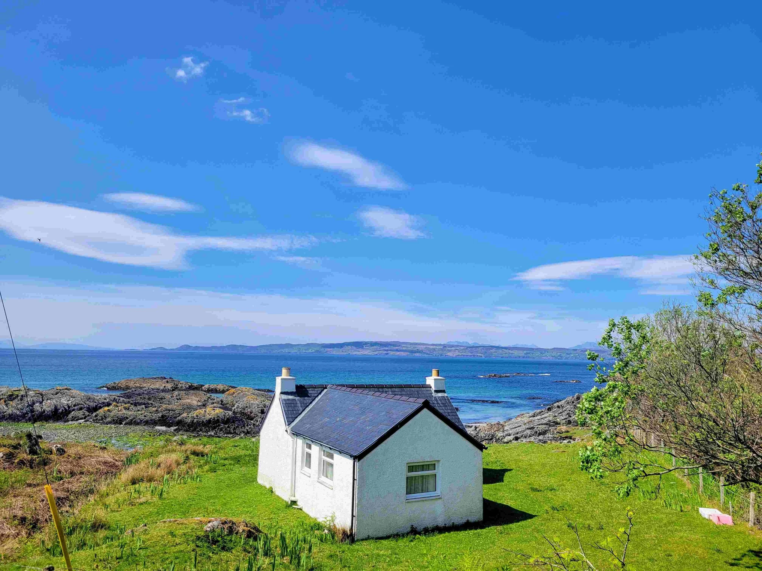 A coastal scene with a white house with a dark roof in the foreground, rocks near the shore, water and distant land across the bay, blue sky with some clouds, and a tree on the right side.