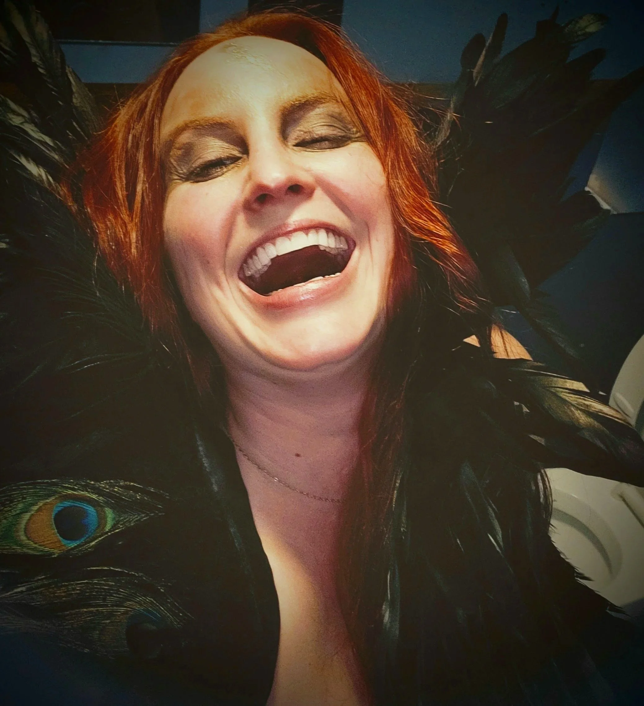 Close-up of a woman with red hair, smiling widely with her eyes closed, adorned with peacock feathers.