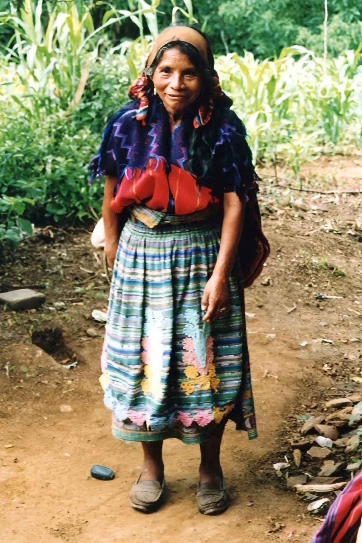 An elderly woman standing outdoors in a garden with green plants in the background. She is wearing a patterned skirt, a colorful shawl, and slip-on shoes, and is smiling at the camera.