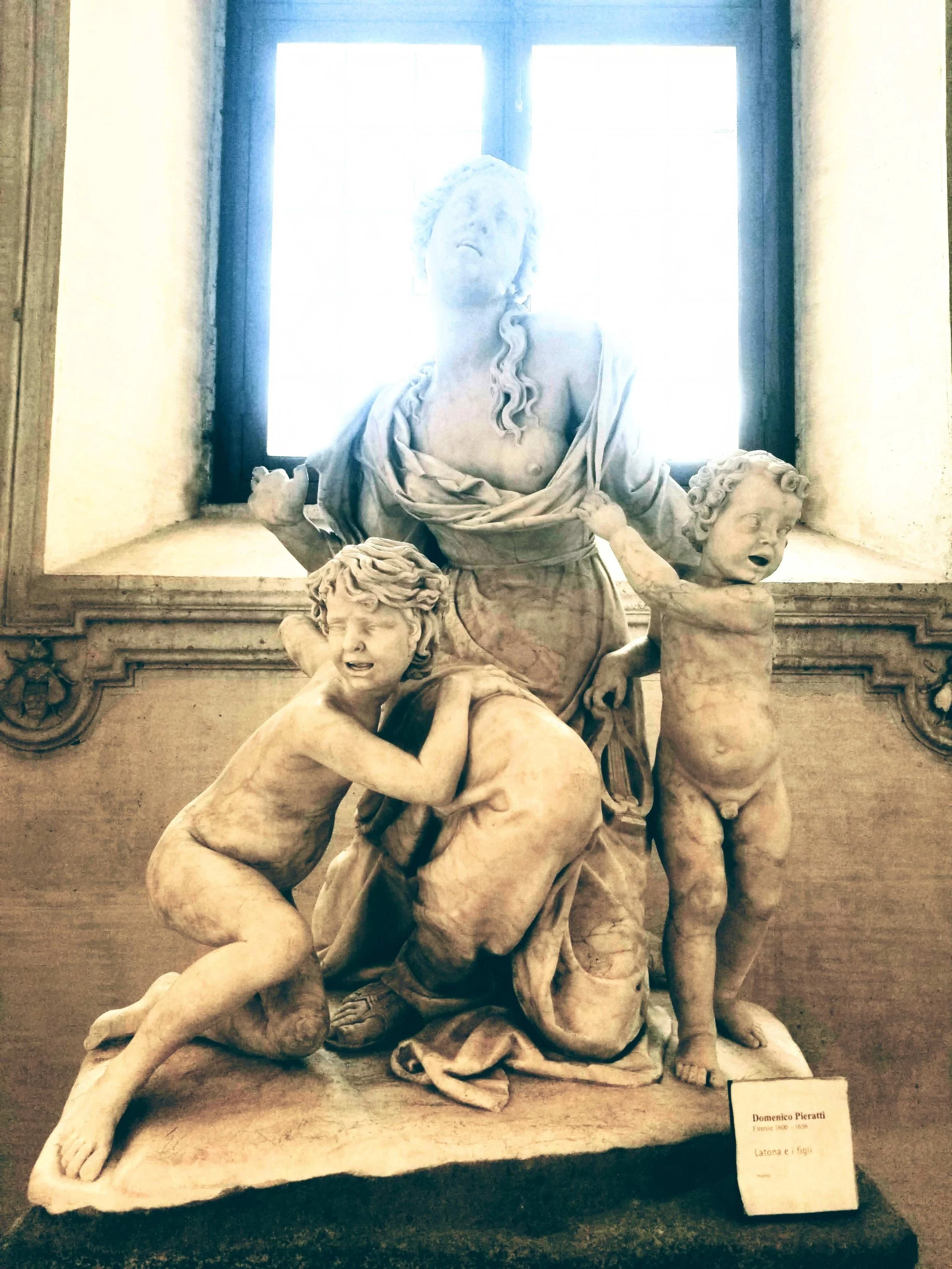 Marble sculpture of a woman with curly hair, holding a child's head with both hands, while another child's head is at her side. The sculpture is displayed in front of a window. A small sign in front of the sculpture reads 'Domenico Pieratti 1601-1656 Latoona e figli'.