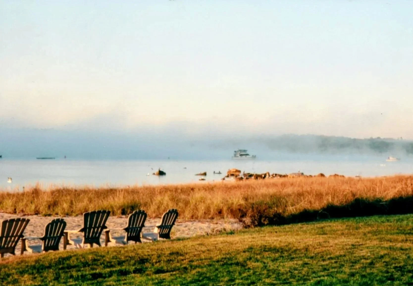 A beach scene at sunset with Adirondack chairs on green grass, a sandy shoreline with rocks, and boats in calm water with a hazy sky in the background.