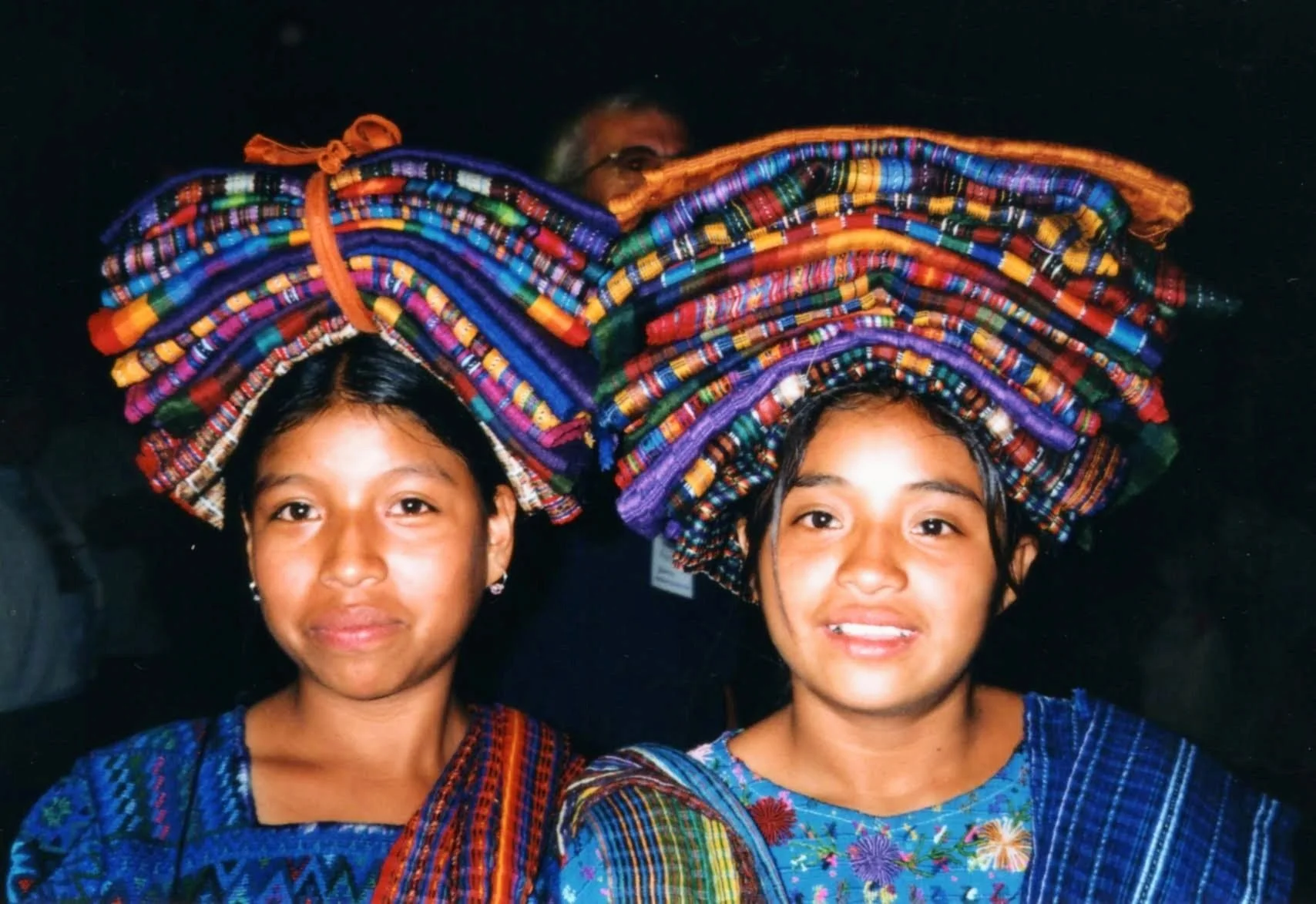Two young girls with traditional woven clothing and large colorful woven headpieces standing against a dark background.