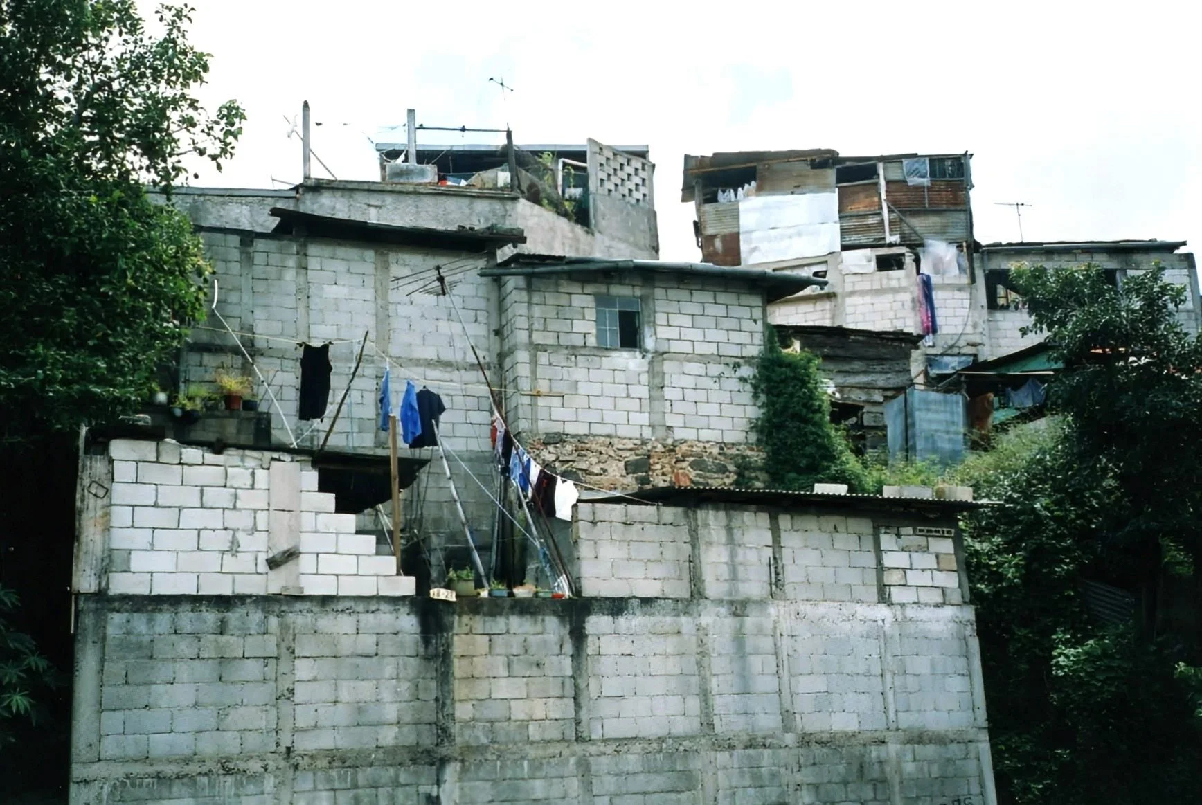 A hillside community with multiple houses made of concrete and corrugated metal, with clothes hanging on lines to dry, surrounded by greenery.