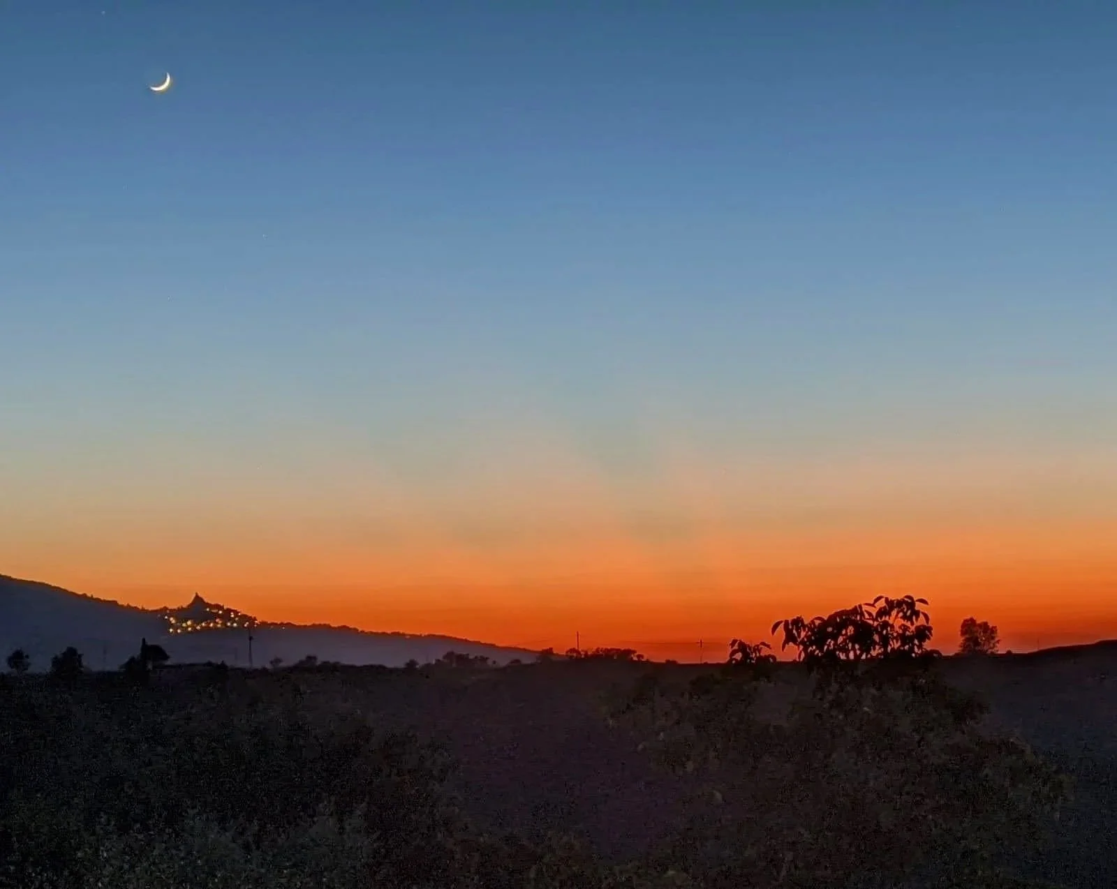Sunset over a landscape with trees and distant hills, a crescent moon in the sky.