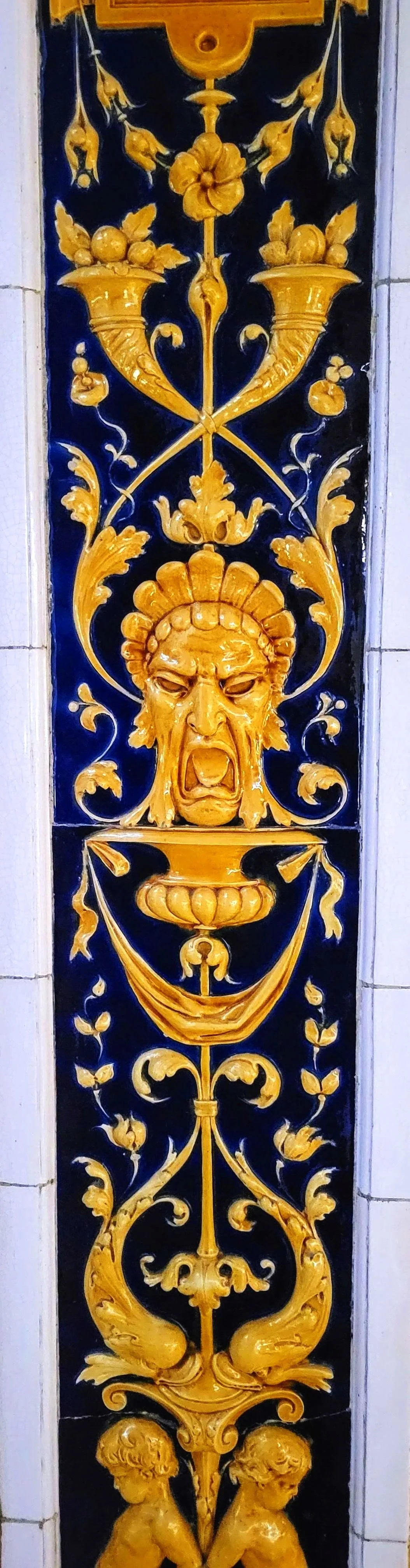 Vertical ceramic tile featuring intricate gold decorative designs on a black background, including a central grotesque face with an open mouth, surrounded by ornate floral and scroll motifs.
