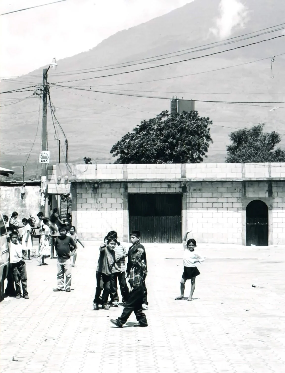 Children playing and standing in a schoolyard or open area in front of a brick building with a large gate, with mountains and power lines in the background.