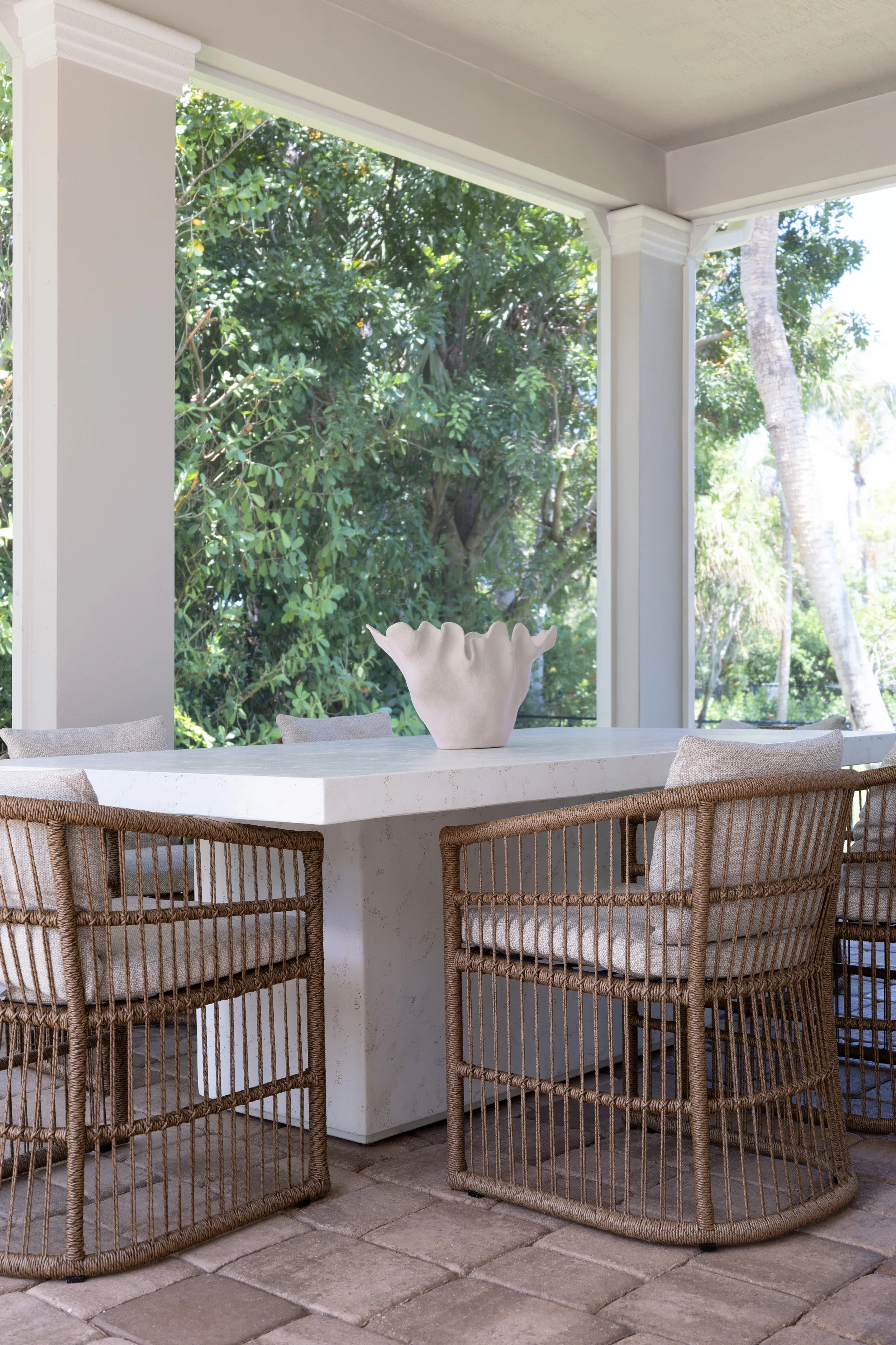 Outdoor dining area with wicker chairs, a white table and sculptural vase on a covered patio surrounded by lush greenery.