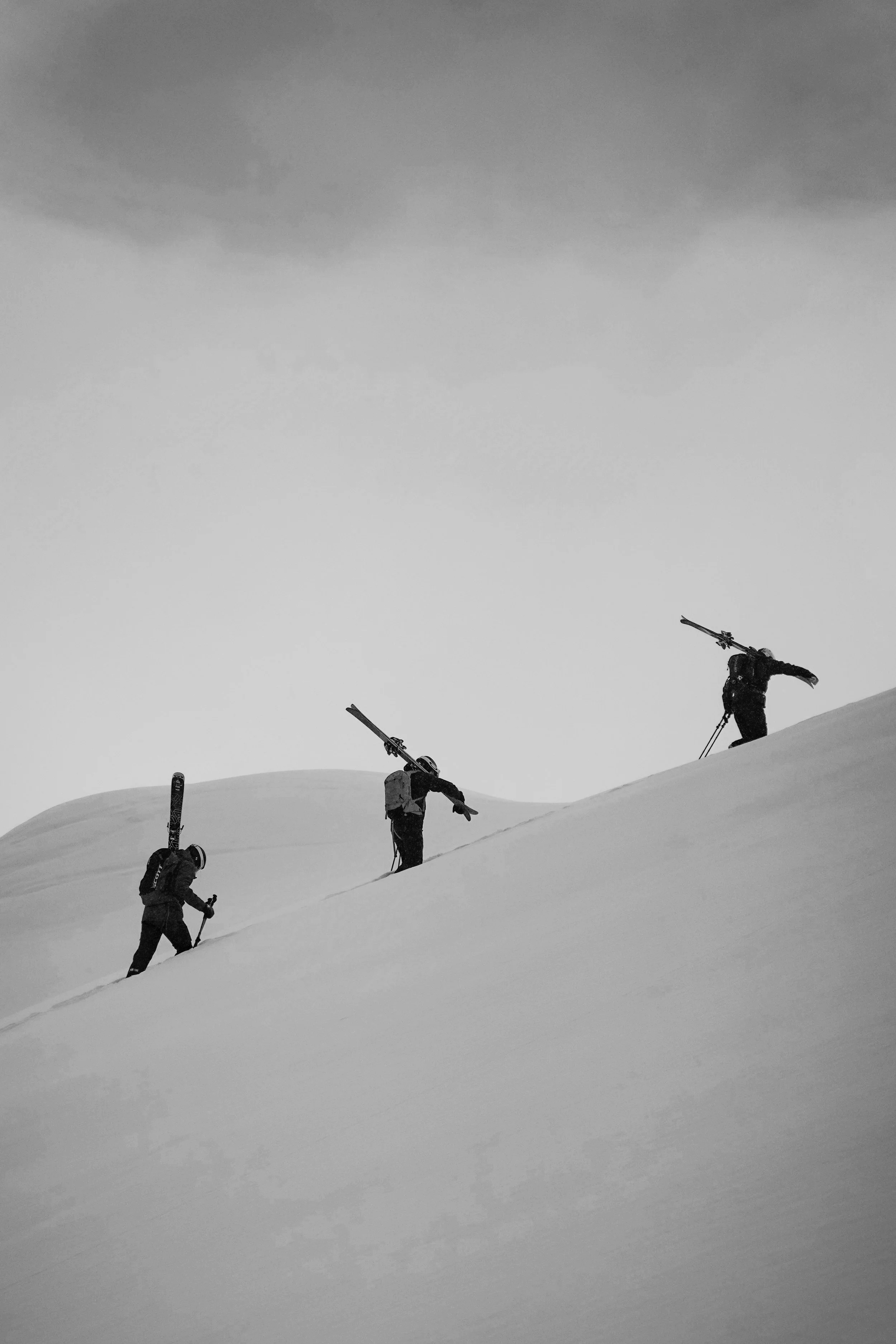Drei Skitourengeher auf einer schneebedeckten Hügellandschaft, einer trägt Skier auf dem Rücken.