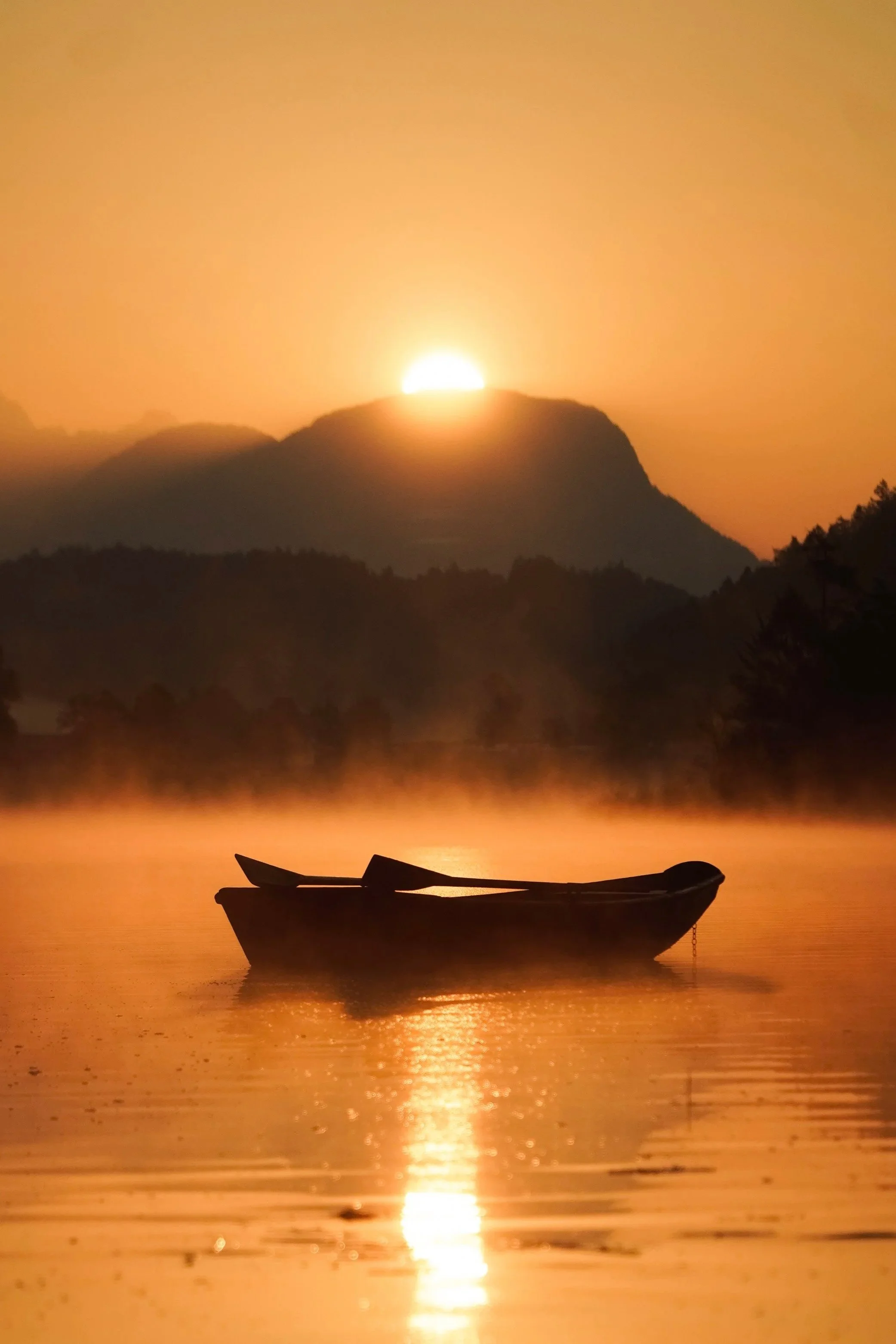 Ein ruhiges Boot schwimmt auf einem See im Sonnenaufgang, mit Bergen im Hintergrund und Nebel über dem Wasser.