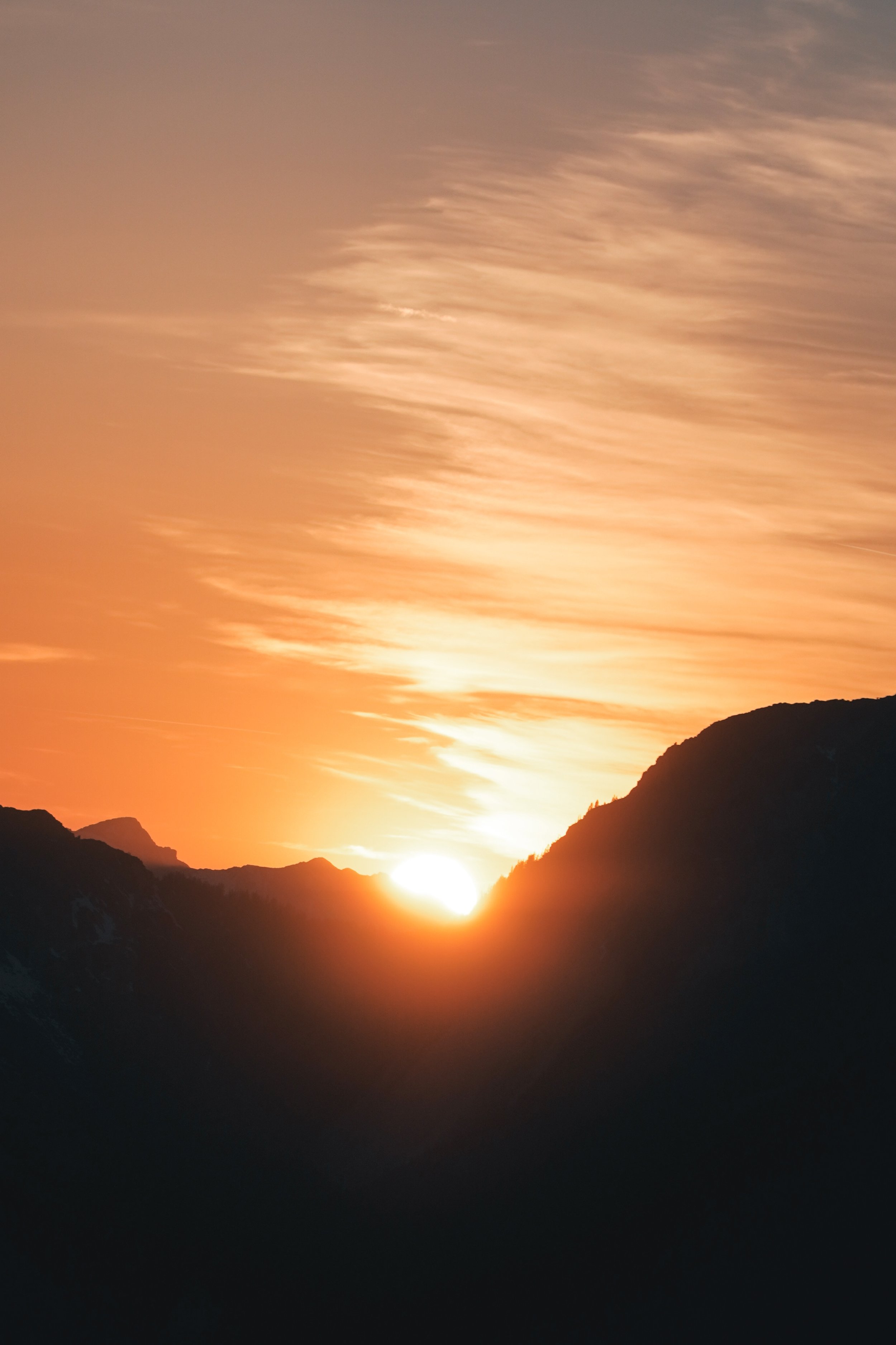Sonnenuntergang hinter Berggipfeln mit orangefarbenem Himmel und sanften Wolken.