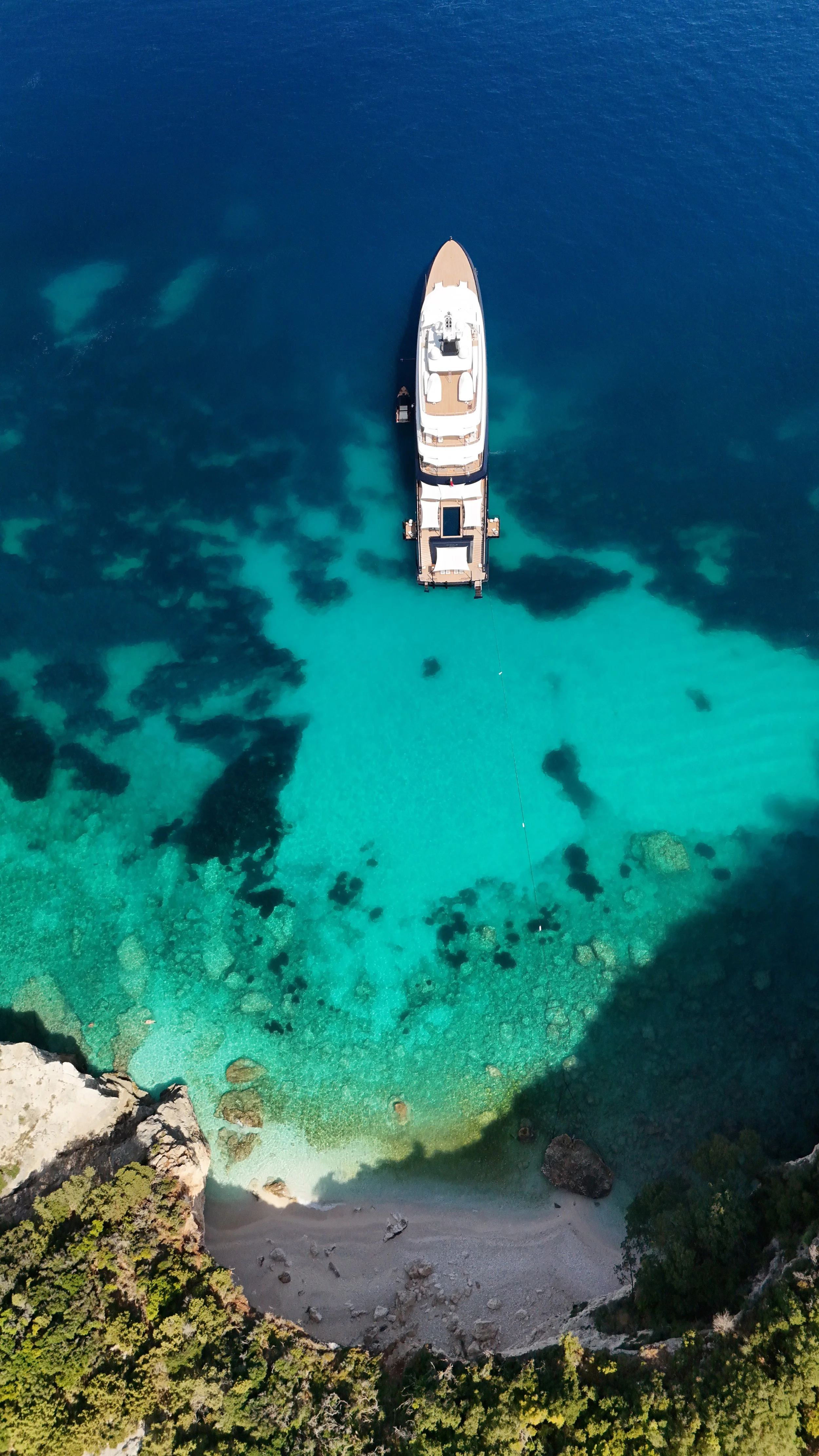 Ein Yachthafen mit einem großen weißen Boot vor türkisfarbenem Meer und einer kleinen Bucht im Hintergrund, umgeben von Felsen und grünen Bäumen.