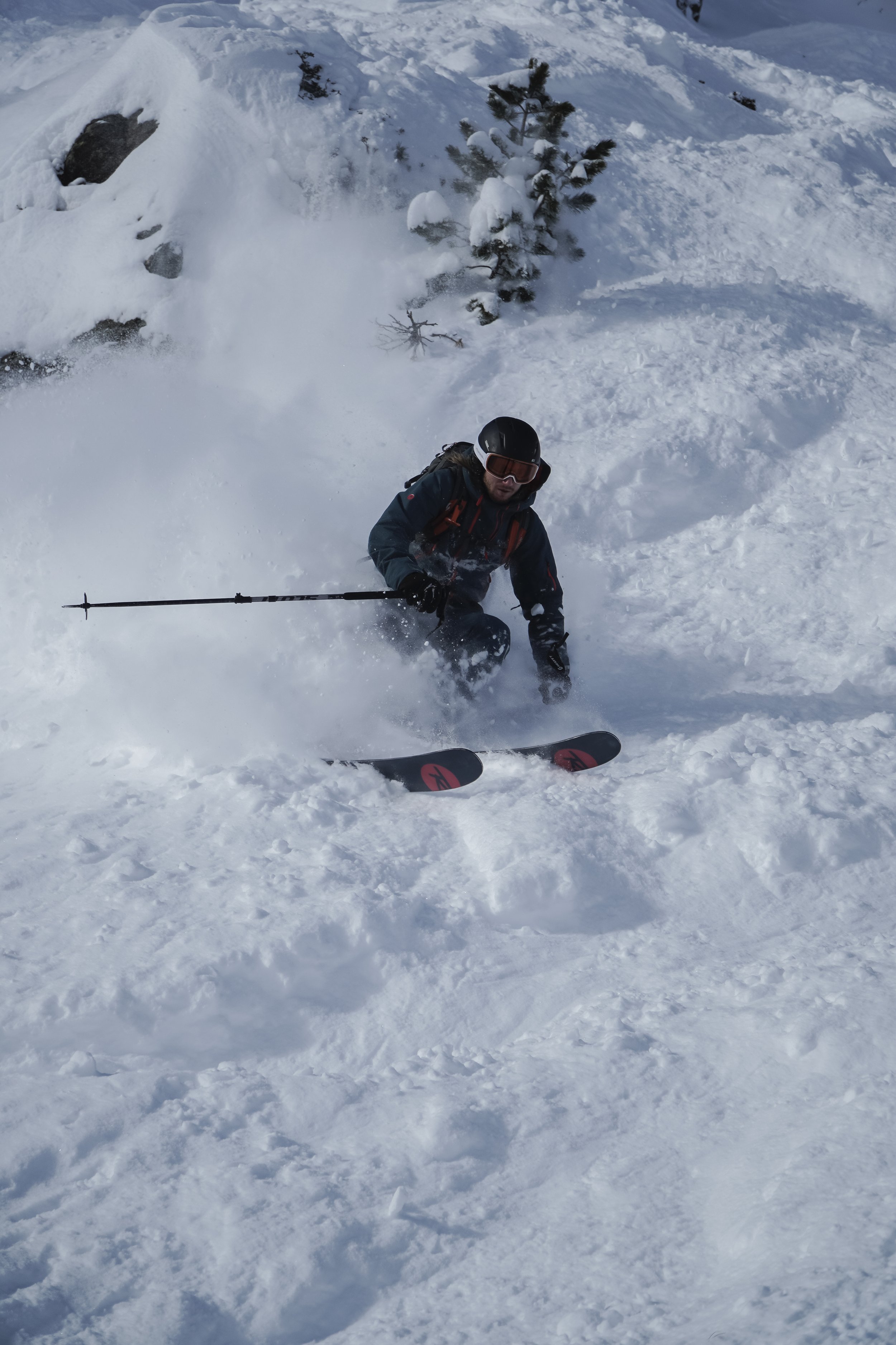 Ein Skifahrer in dunkler Kleidung, mit Helm und Skibrille, fährt in tiefem Schnee in einer winterlichen Berglandschaft.
