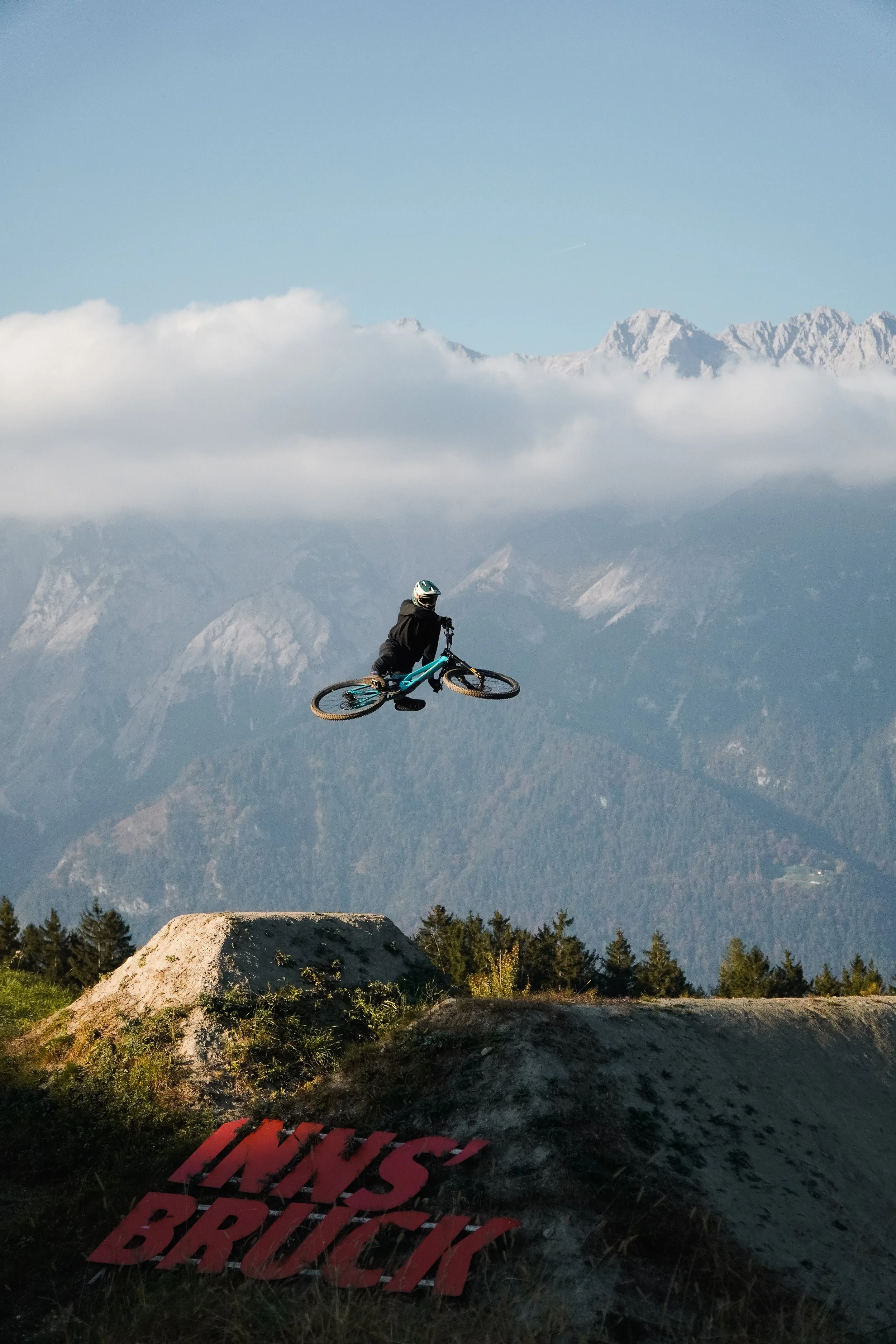 Mountainbiker springt über einen Sprung in einem Outdoor-Park mit Bergen im Hintergrund. Es gibt einen roten Schriftzug, der 'LINK'S BRUCK' sagt.