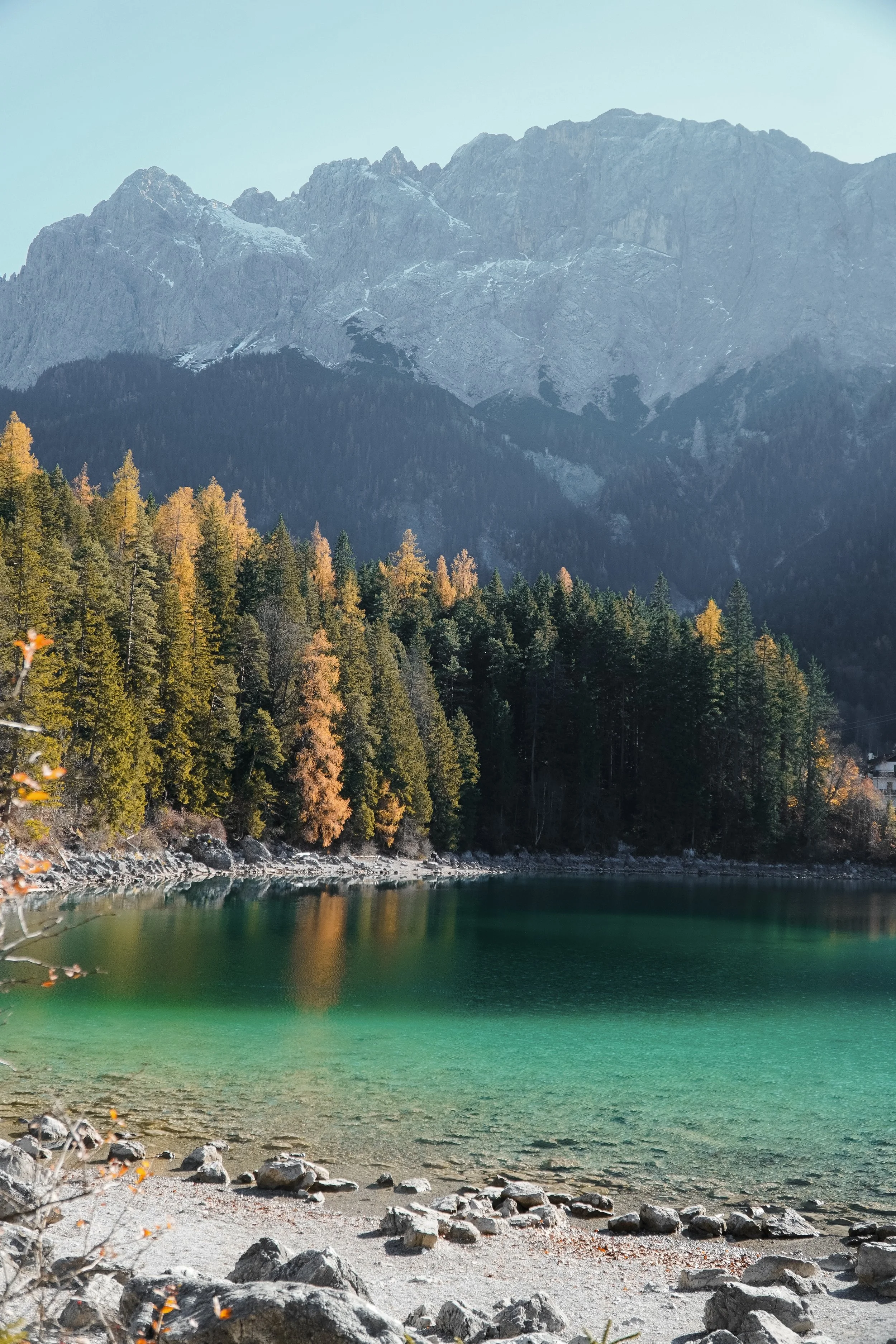Bergsee mit grüner Wasser, umgeben von steiniger Küste, im Hintergrund ein bewaldeter Hang mit Herbstbäumen und hohe bergige Gipfel.