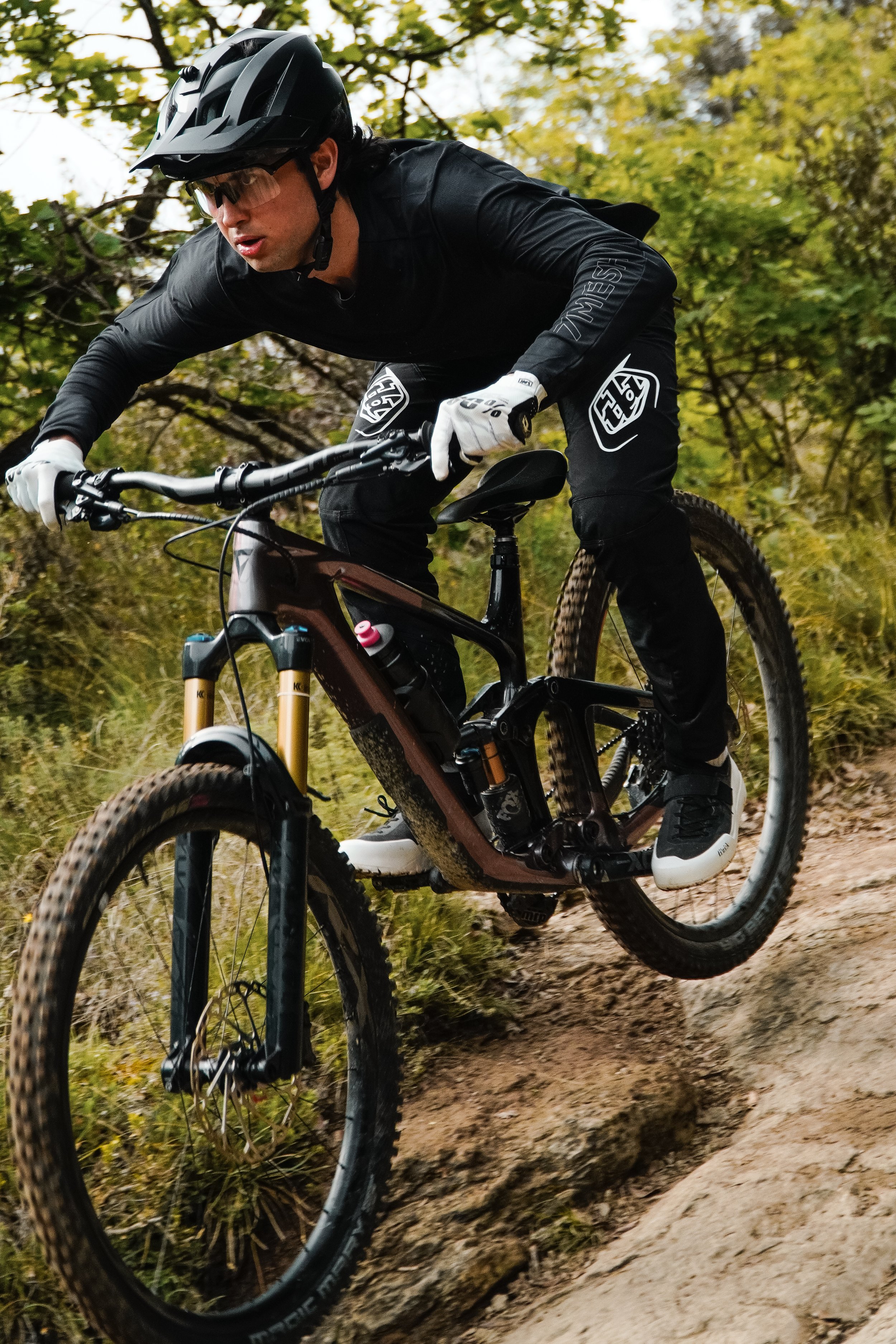 Ein Mountainbike-Fahrer trägt einen schwarzen Helm und schwarze Kleidung, fährt auf einem unbefestigten, steinigen Trail in einen bewaldeten Bereich.