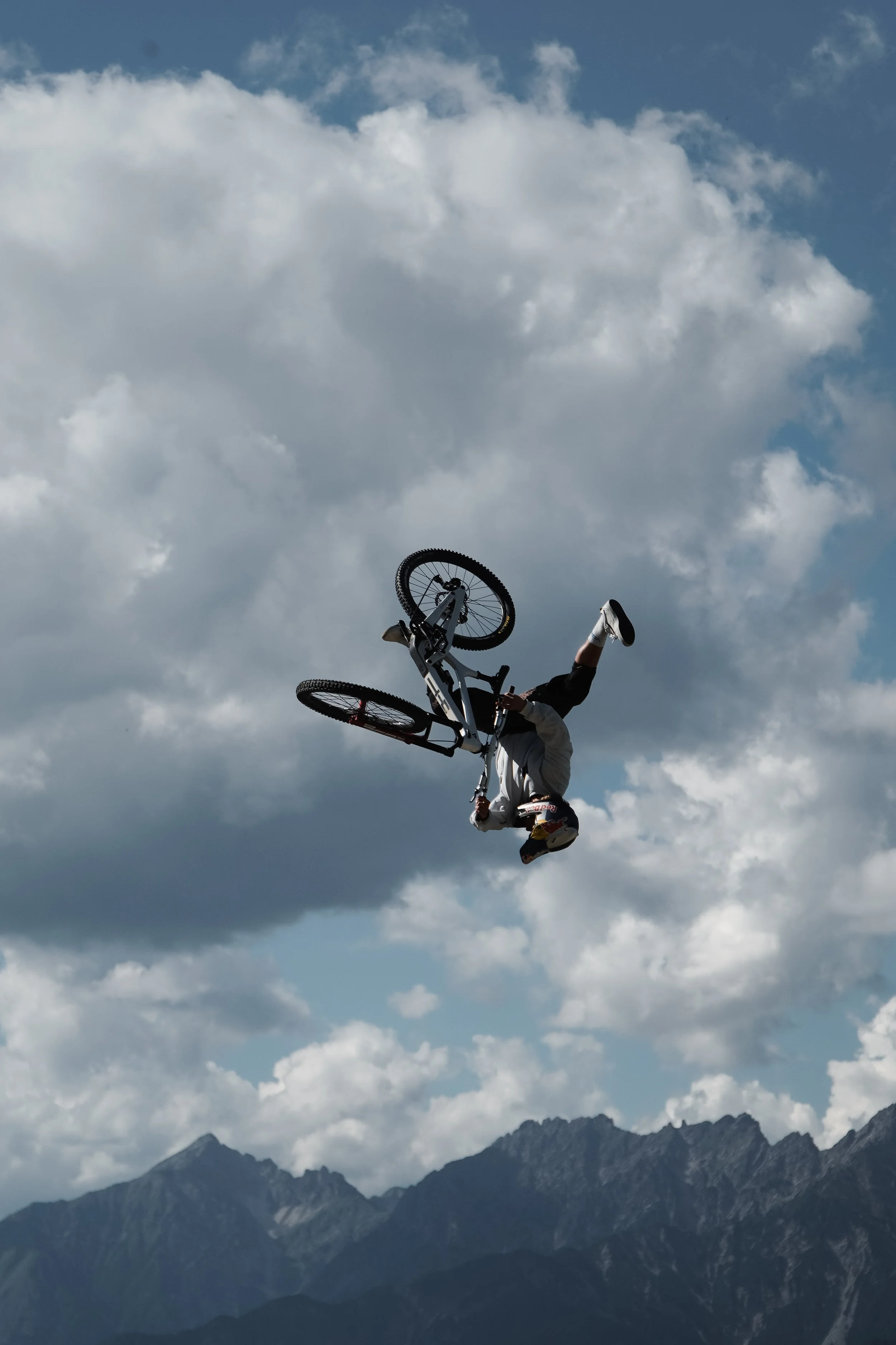 Mountainbiker in der Luft während eines Tricks mit einem Himmel voller Wolken und Berggipfeln im Hintergrund.