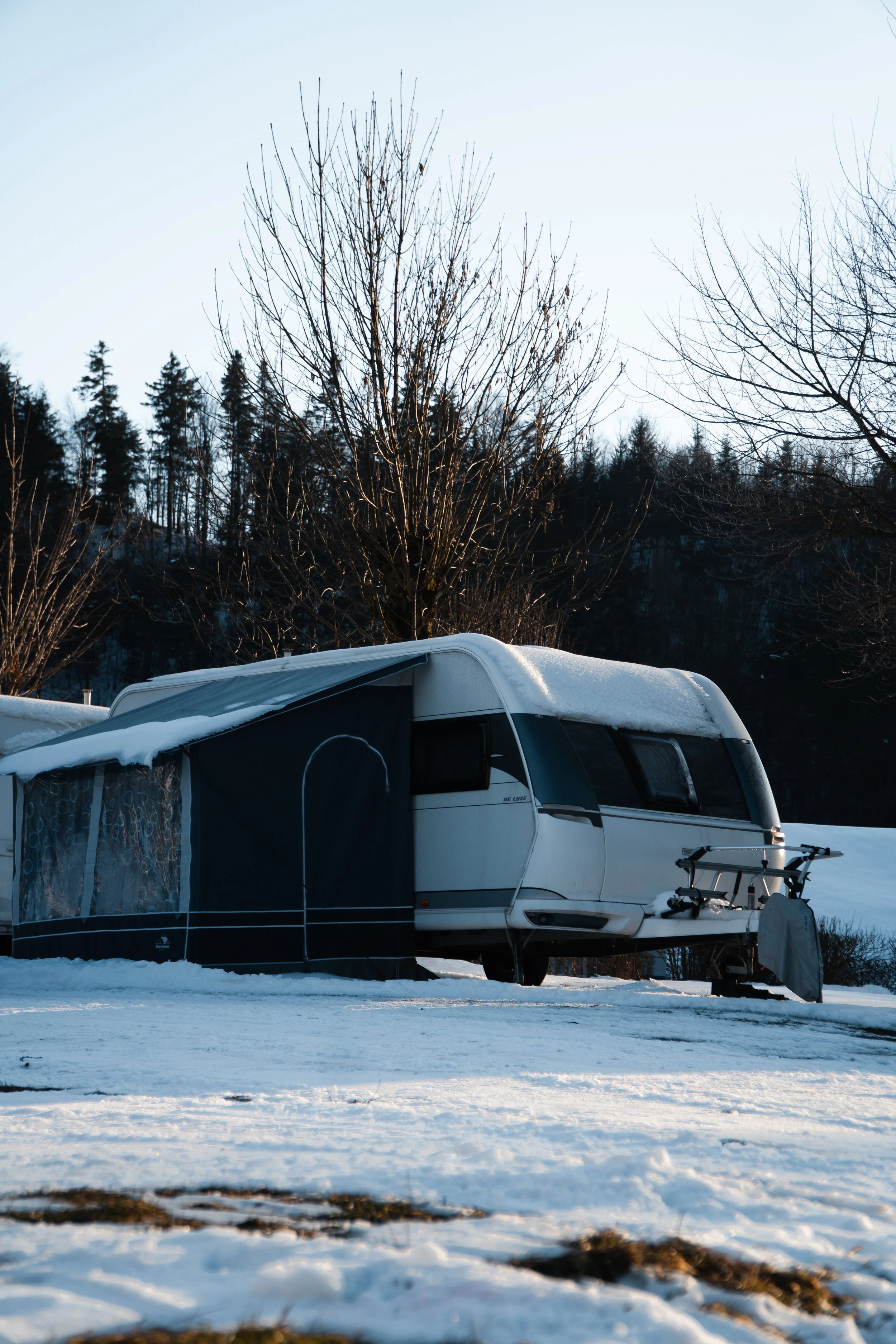 Ein Wohnwagen steht im Schnee vor einem Baum mit kahlen Ästen, im Hintergrund Wald und Berge, es ist Winter.