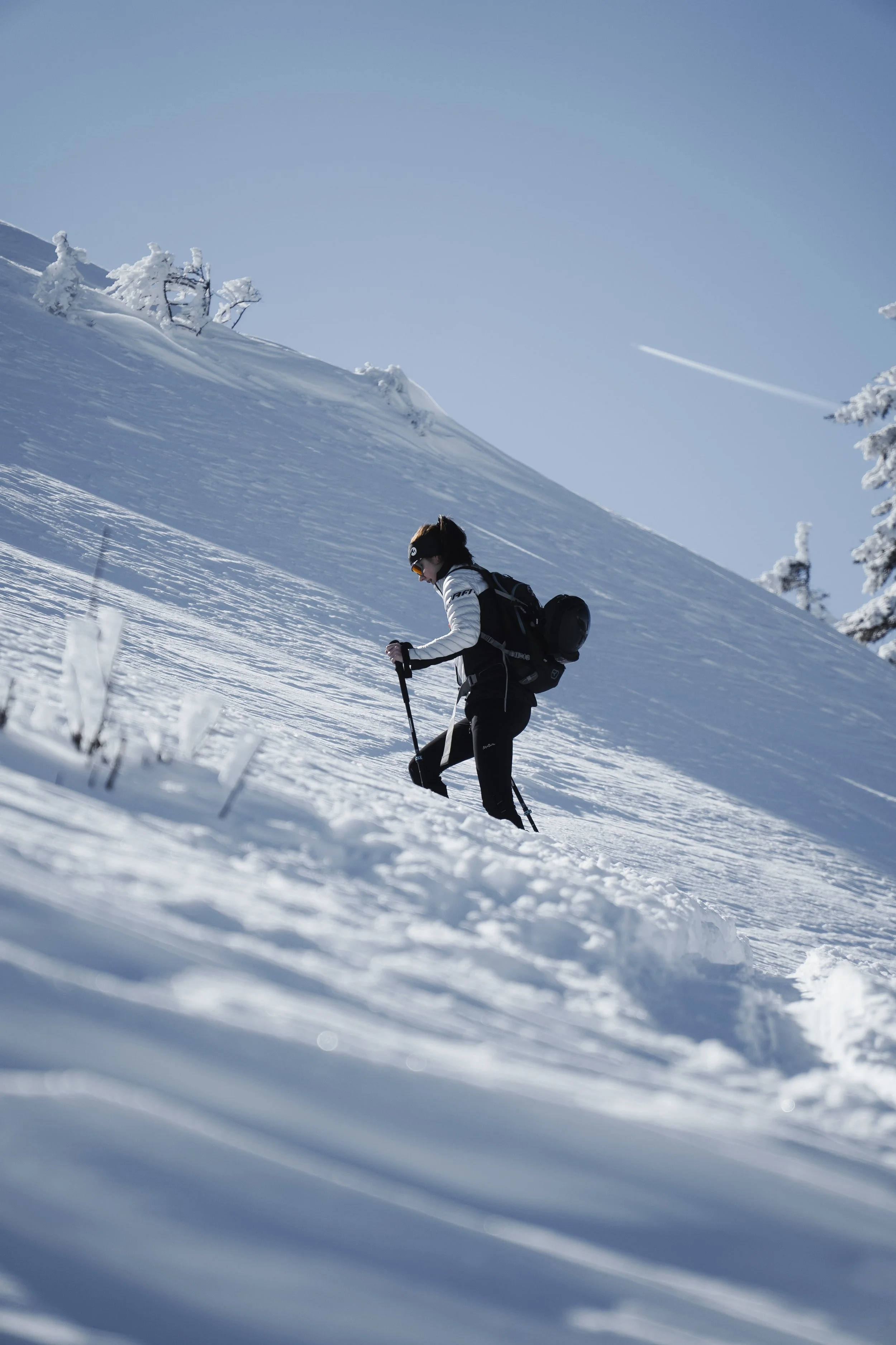 Skifahrerin in Winterlandschaft, die einen Hang hinaufsteigt, mit Schneeschuhen und Rucksack, bei klarem Himmel.