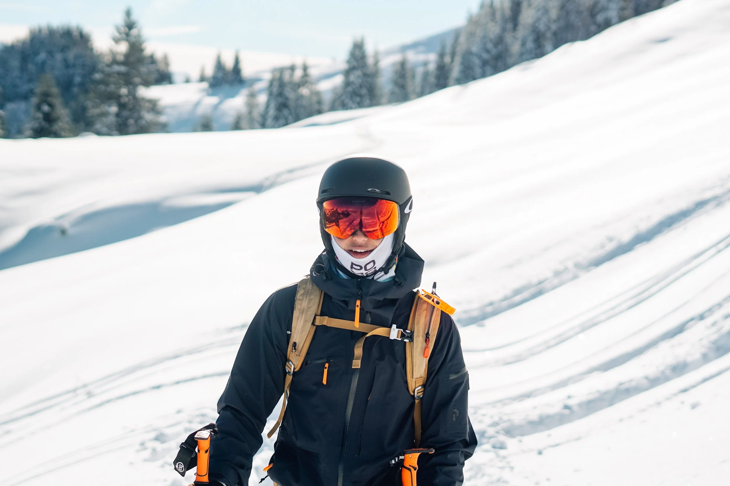 Ein Skifahrer in schwarzer Skibekleidung mit orangefarbenen Akzenten, schwarzer Helm, rotem Skibrillenglas und einem Rucksack, steht in einer verschneiten Winterlandschaft mit Bäumen im Hintergrund.
