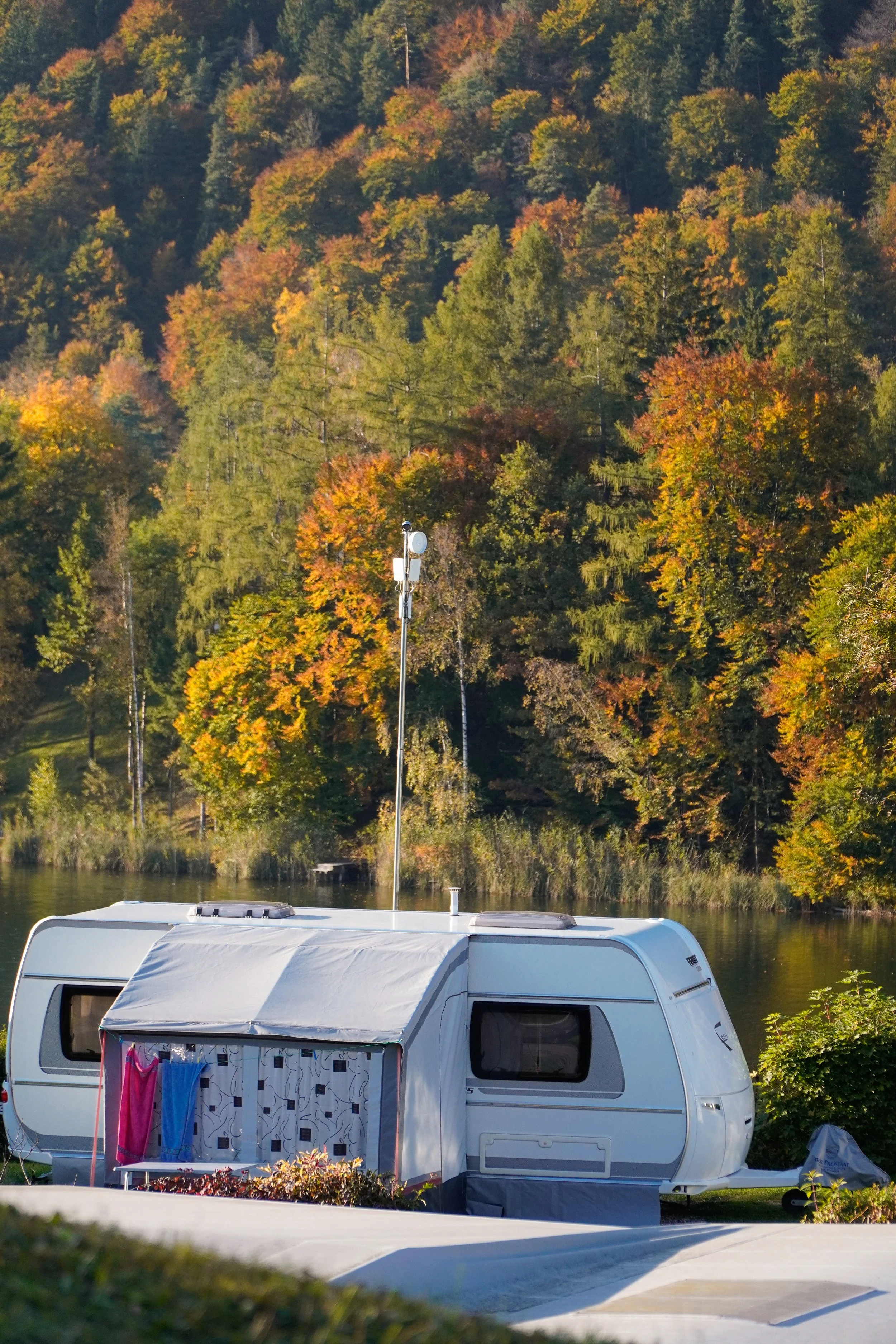 Ein Wohnwagen steht an einem See, im Hintergrund sind Bäume mit buntem Herbstlaub, es ist ein sonniger Tag.