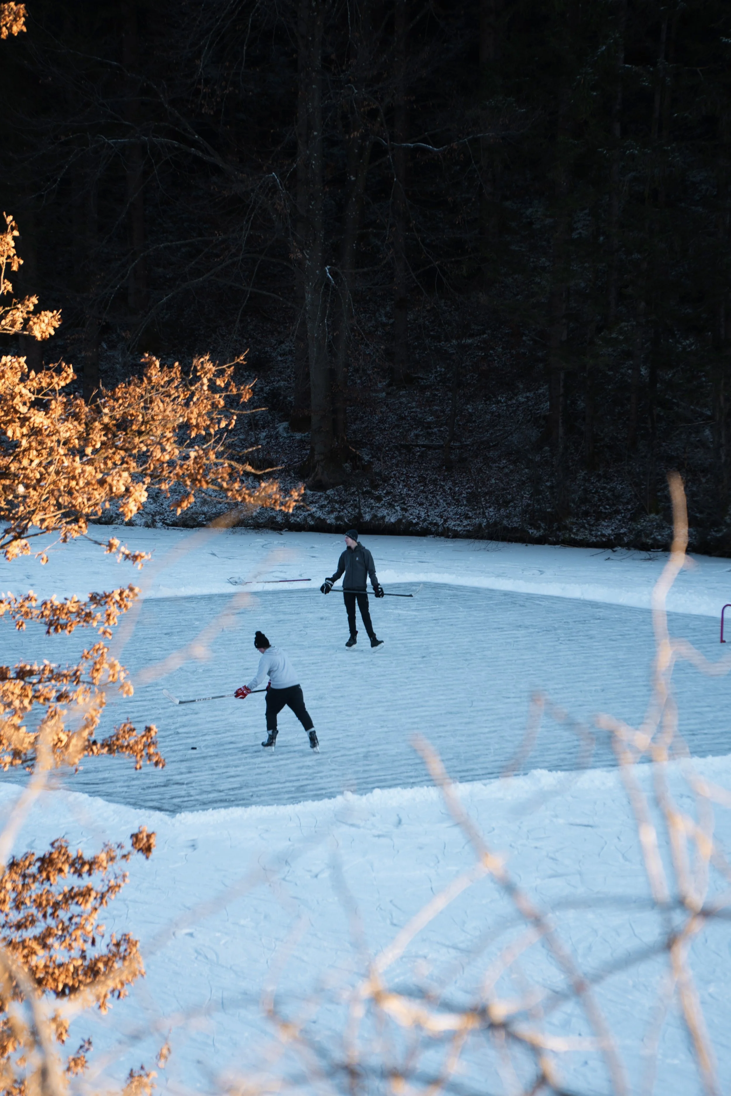 Zwei Personen beim Eishockeyspielen auf einem gefrorenen See im Winter, umgeben von Bäumen, bei Sonnenuntergang.