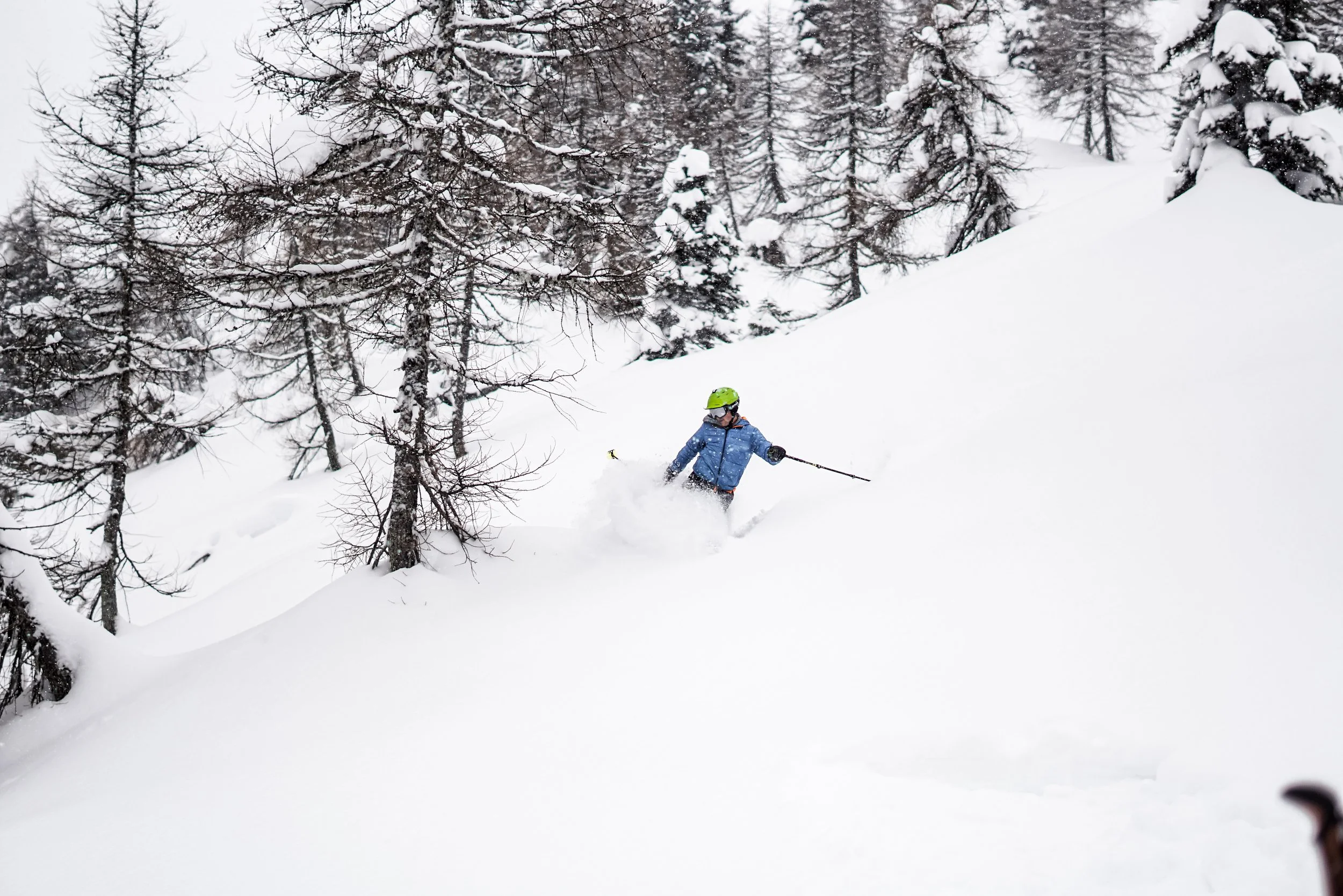 Skifahrer in blauer Jacke und grünem Helm fährt durch tiefen Schnee in einem verschneiten Wald.