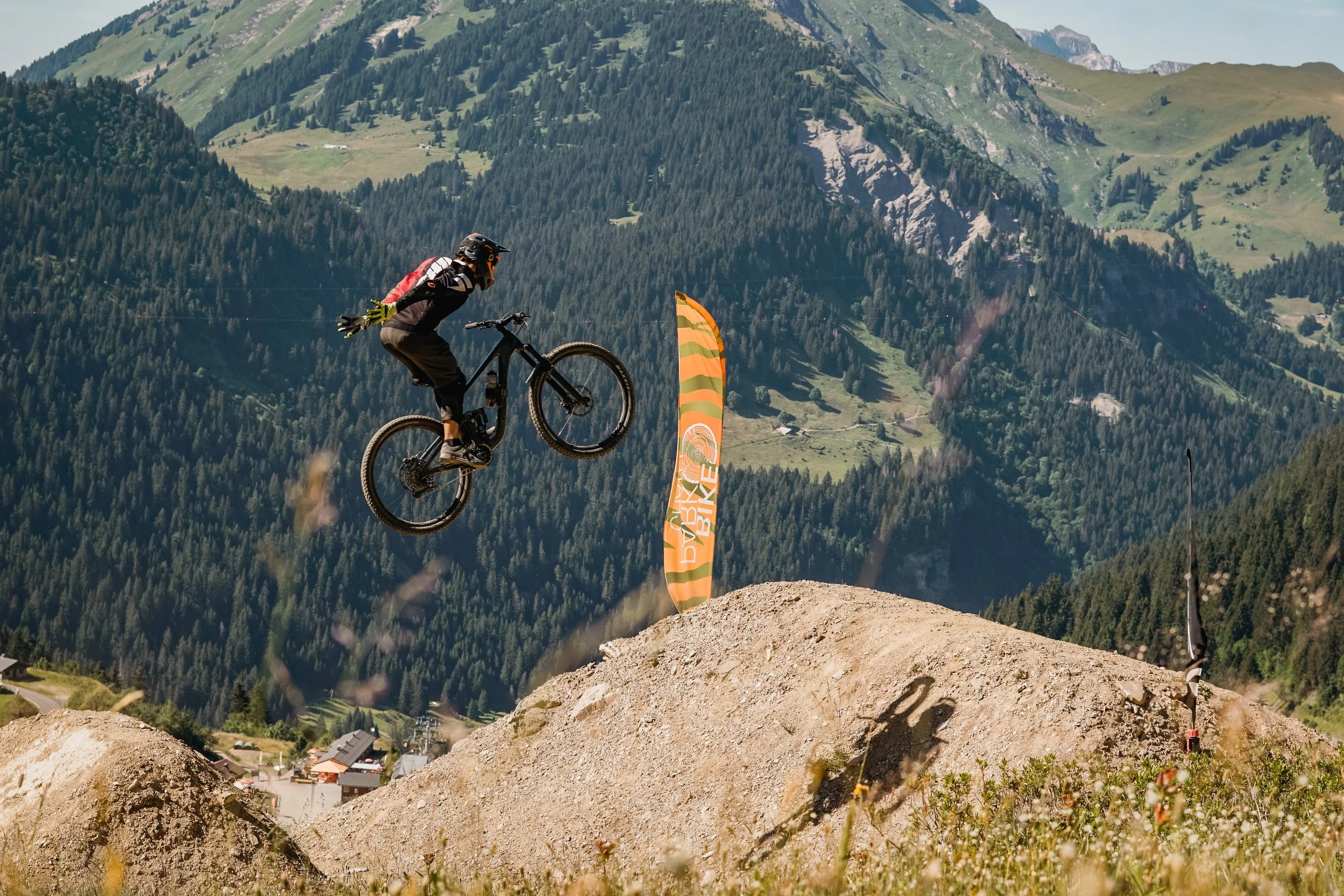 Mountainbiker in der Luft bei einer Sprungaktion auf einer Dirtbike-Strecke in einer Bergregion mit Wäldern im Hintergrund.