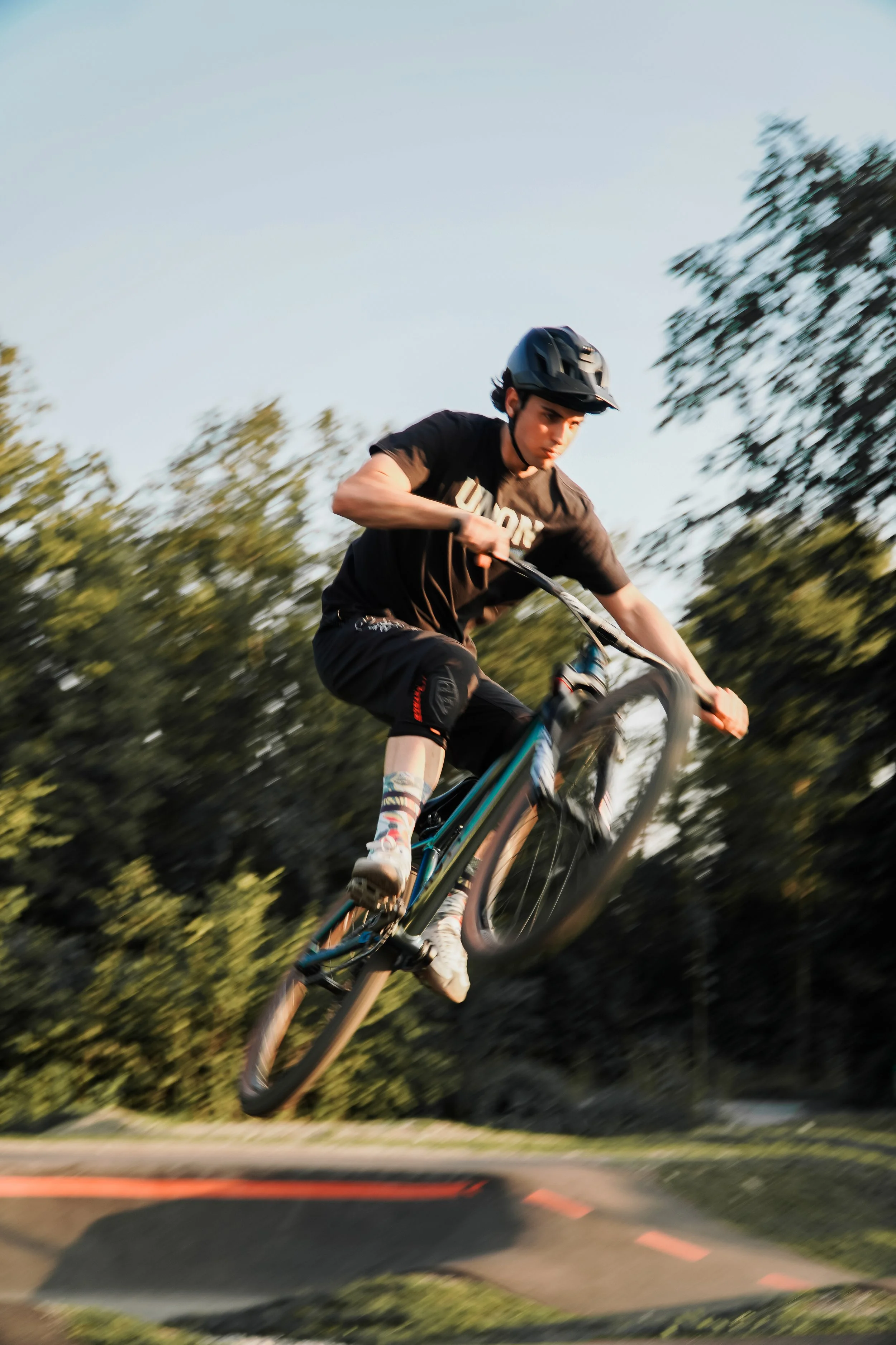 Junge beim Mountainbiken in der Luft auf einem Dirt-Sprung, umgeben von Bäumen, bei Sonnenuntergang.