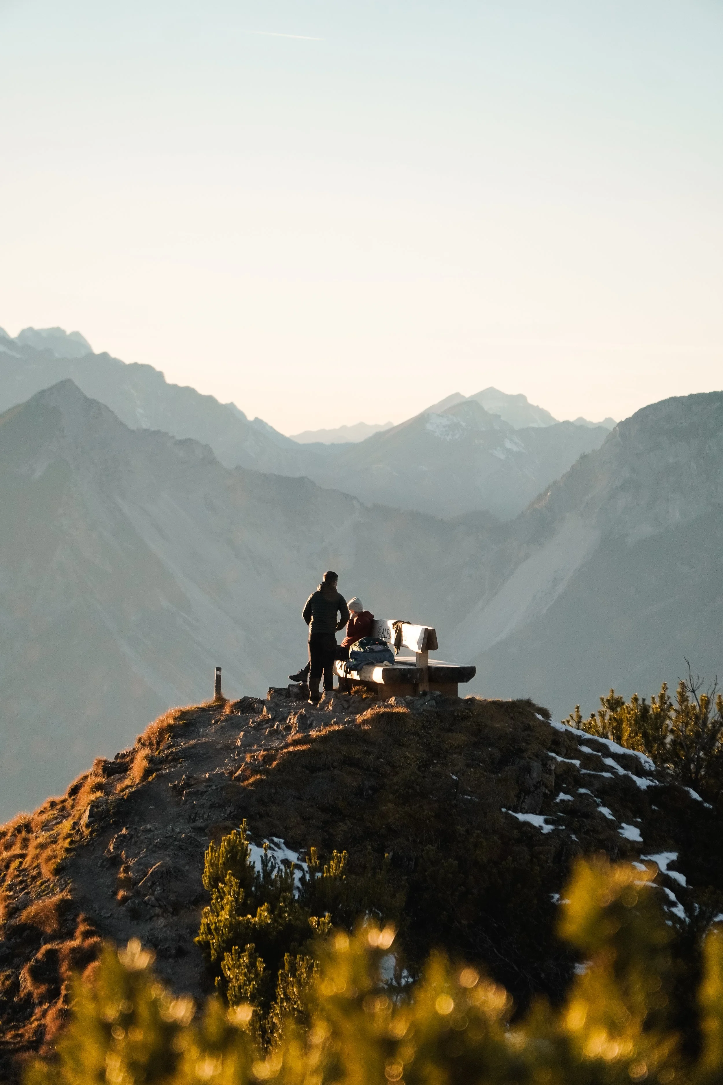 Zwei Personen sitzen auf einer Bank auf einem Berggipfel bei Sonnenuntergang, im Hintergrund sind hohe Berge sichtbar.