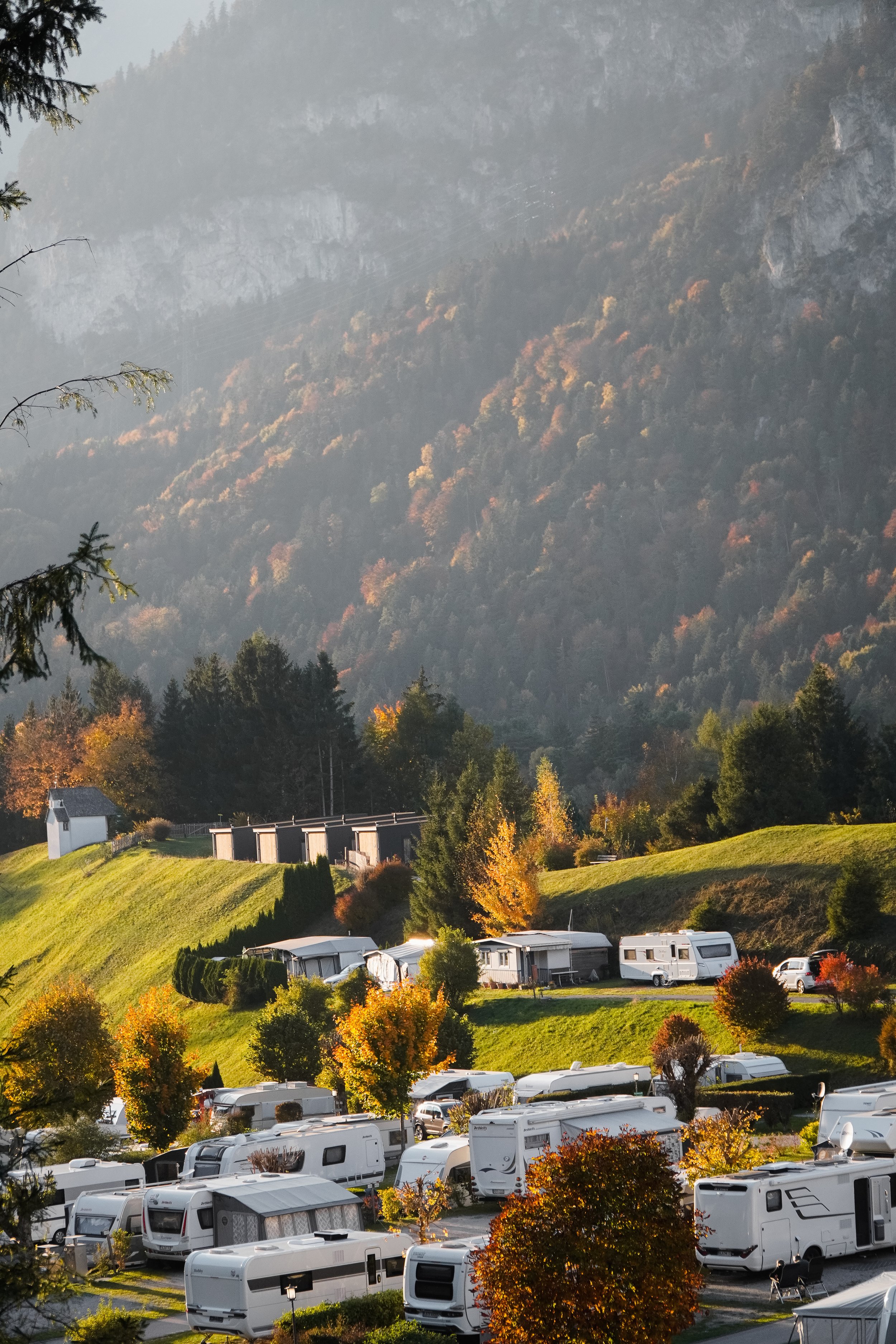 Ein Campingplatz in einer bergigen Landschaft mit Wohnmobilen, Bäumen mit Herbstfärbung und grünen Hügeln im Hintergrund.