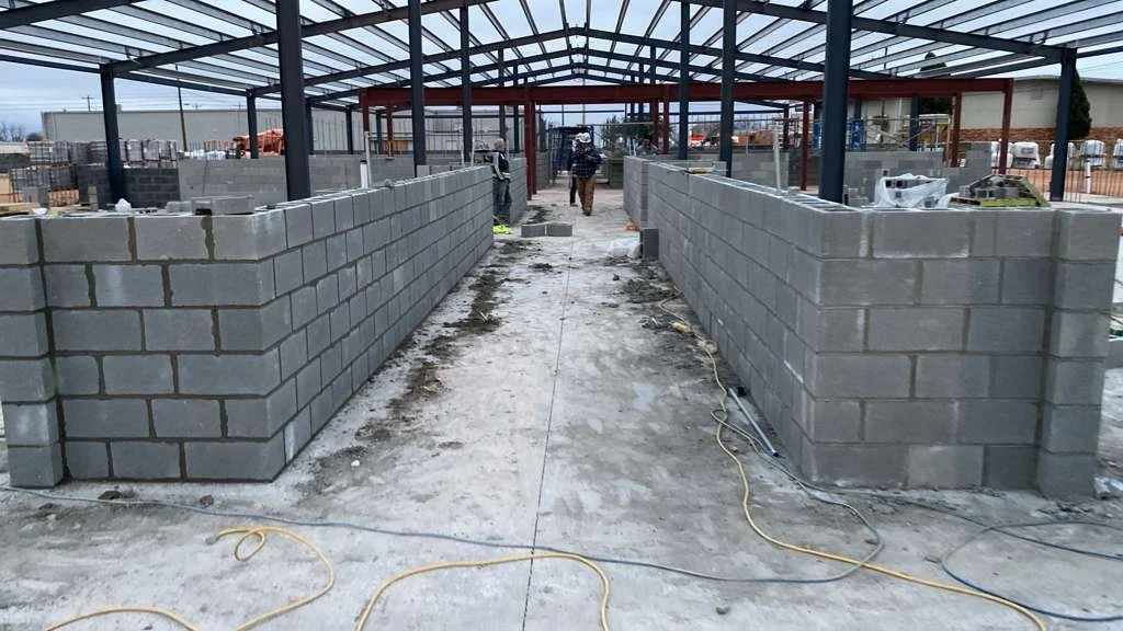 Construction site with gray cinder block walls and steel framework, workers in the background, concrete ground with scattered cords and tools.
