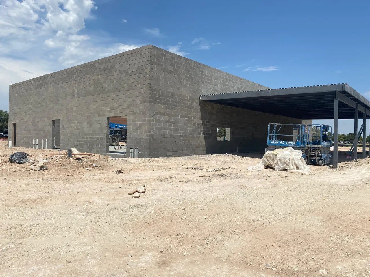 Under construction building with gray cinder block walls, a covered area on the right, and construction equipment on site. The ground is dirt and the sky is partly cloudy.