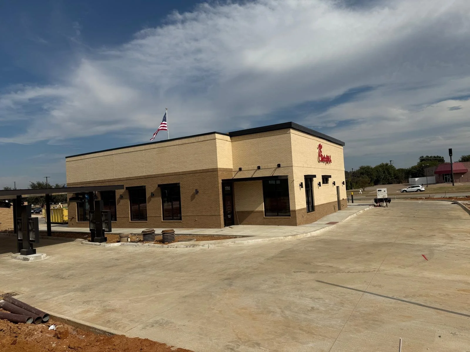 A Chick-fil-A restaurant under construction with a beige brick exterior, American flag on top, and a parking lot in front, with a few cars and a partly cloudy sky.