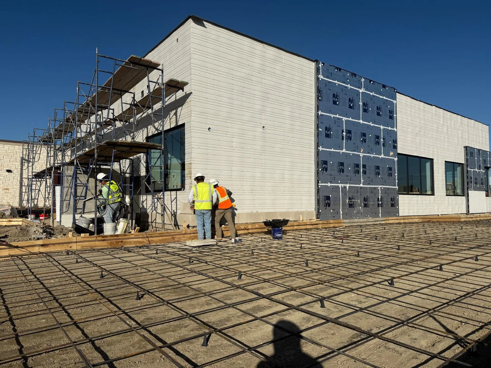 Construction workers in safety vests and helmets working on building exterior with scaffolding and rebar on ground.