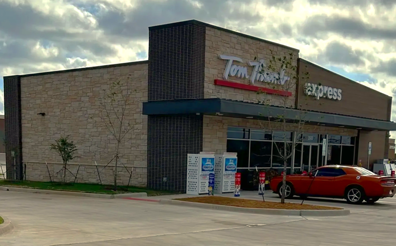 Exterior of a Tom Thumb Express convenience store with a parked orange car in front, some small trees, and two vending machines outside.