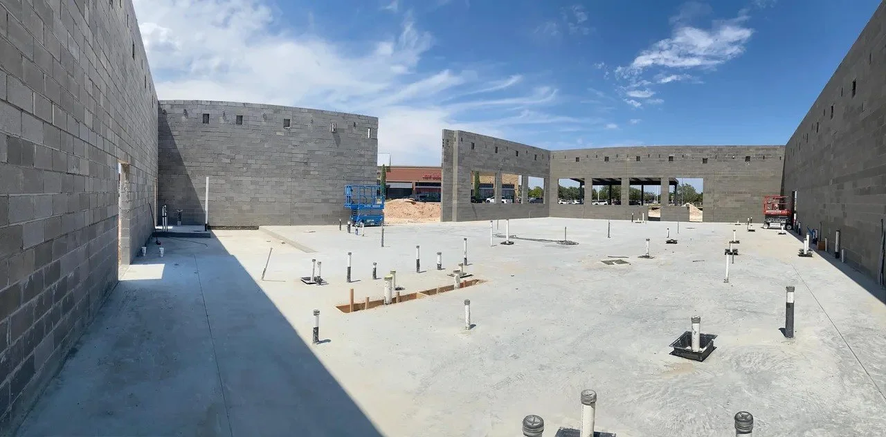 Construction site with a concrete foundation, cinder block walls, and metal pipes emerging from the ground under a blue sky with scattered clouds.