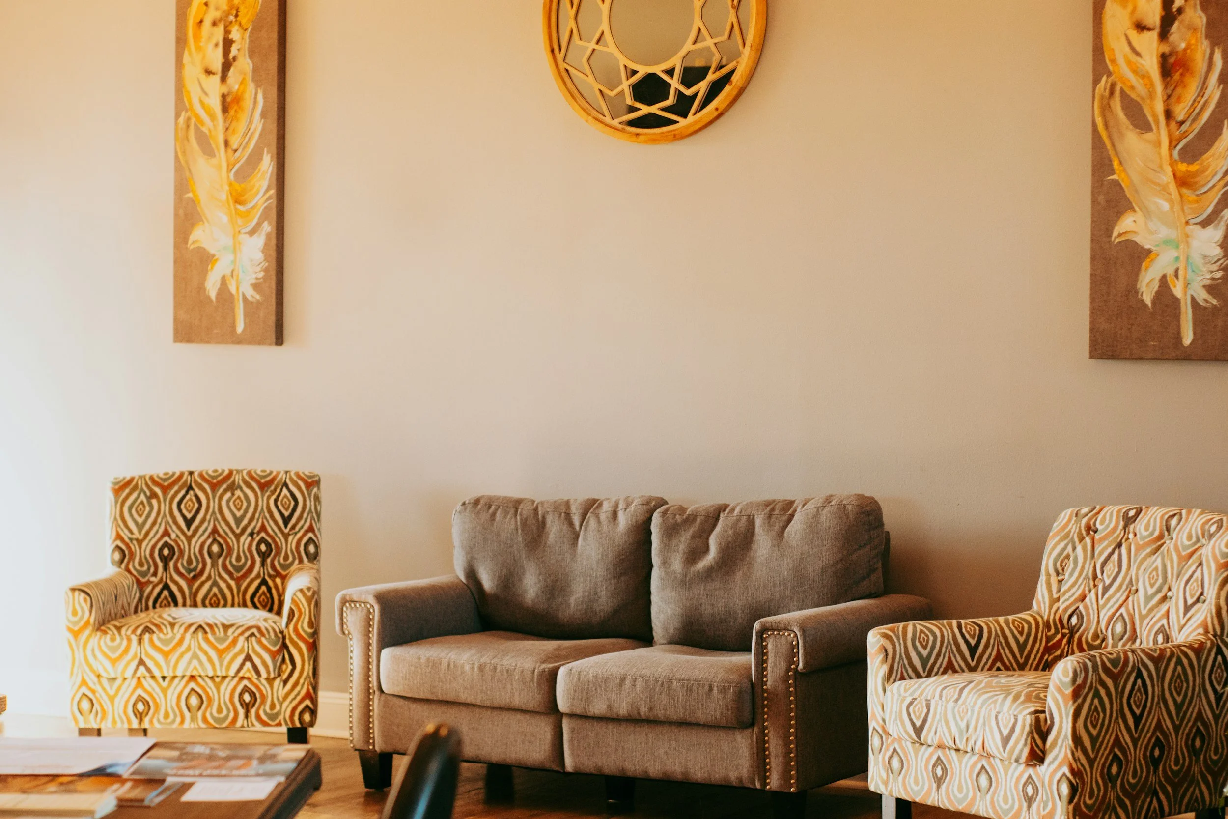 Therapy office with beige sofa, two patterned armchairs, two wall art pieces with feathers, and decorative mirror on the wall. Counseling in Cincinnati, OH offers warmth, peace, love, acceptance, emotional safety, support, a safe space to be yourself