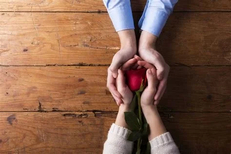 Zwei Hände halten gemeinsam eine rote Rose über einem Holztisch.