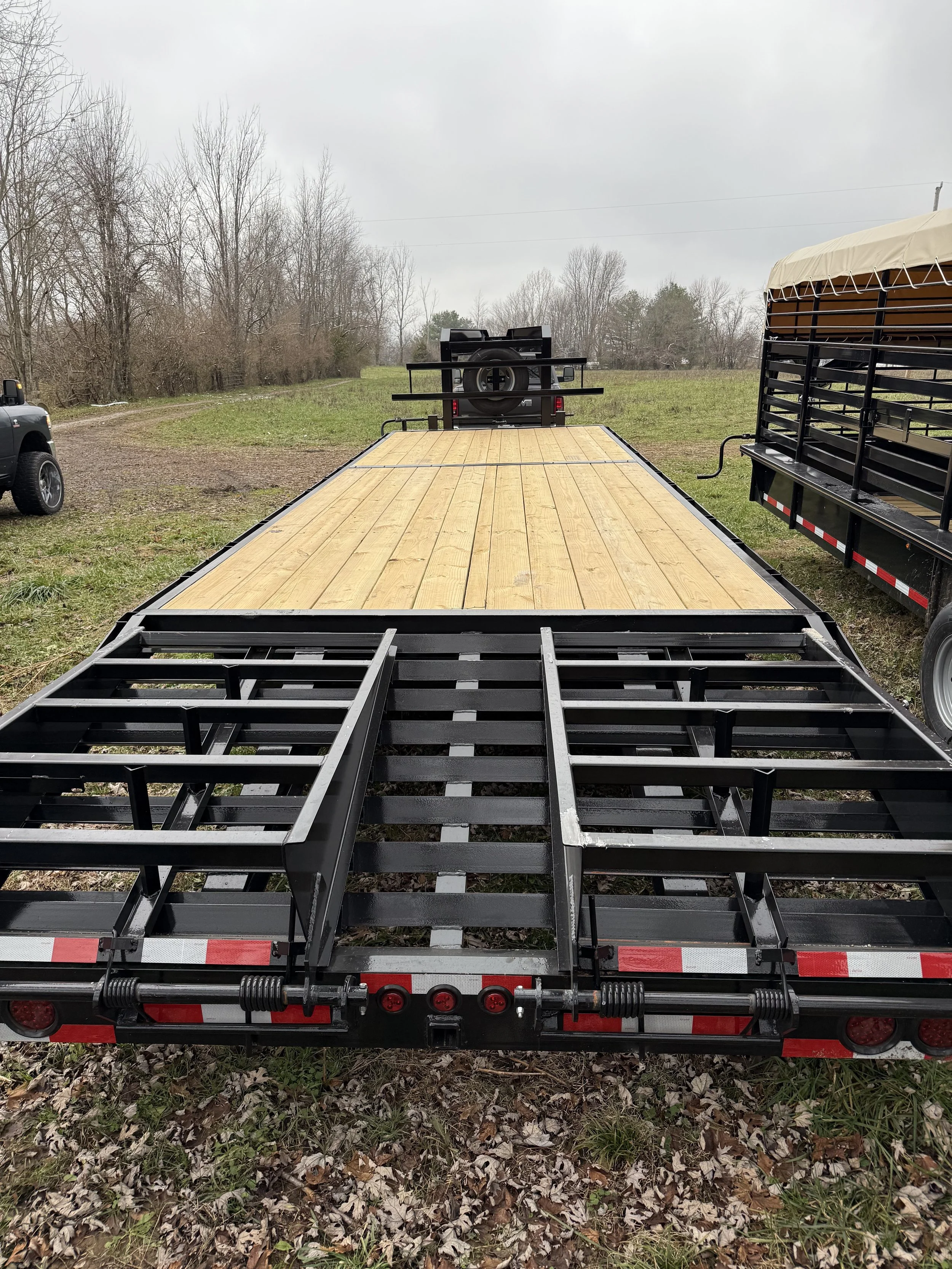 A flatbed trailer with a wooden deck, parked on grass with leaf litter, with another trailer and a vehicle in the background under a cloudy sky.