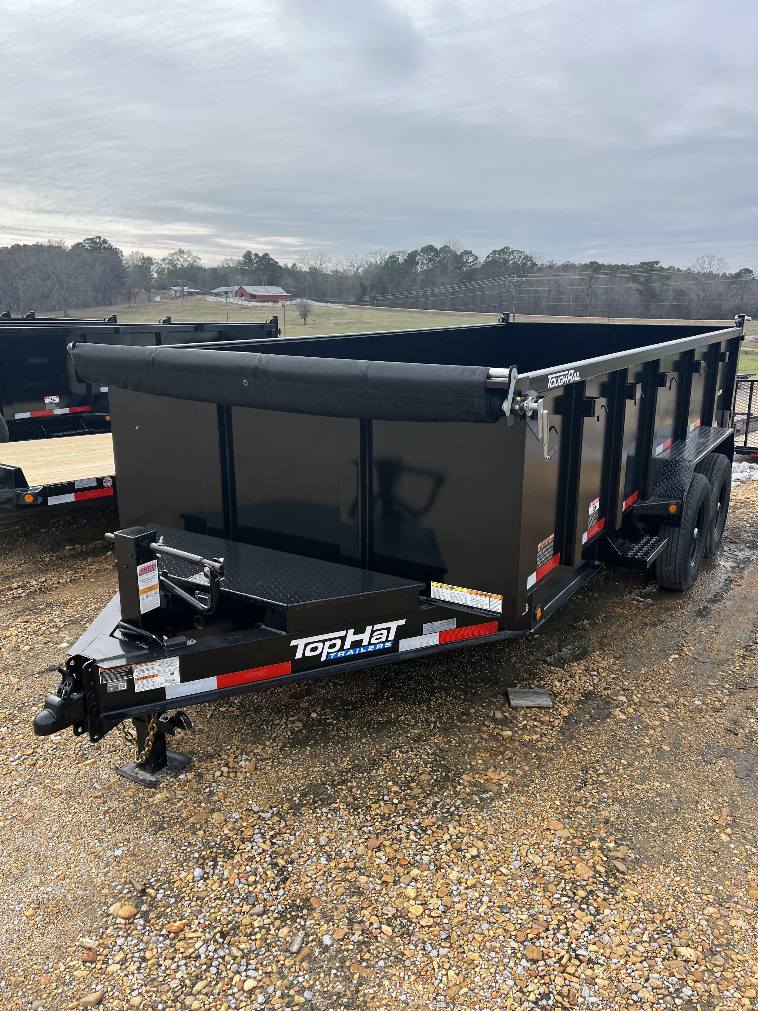 Black utility trailer with a metal frame and wooden flooring, parked on gravel, with a hitch at the front for attaching to a vehicle, outdoors under a cloudy sky.