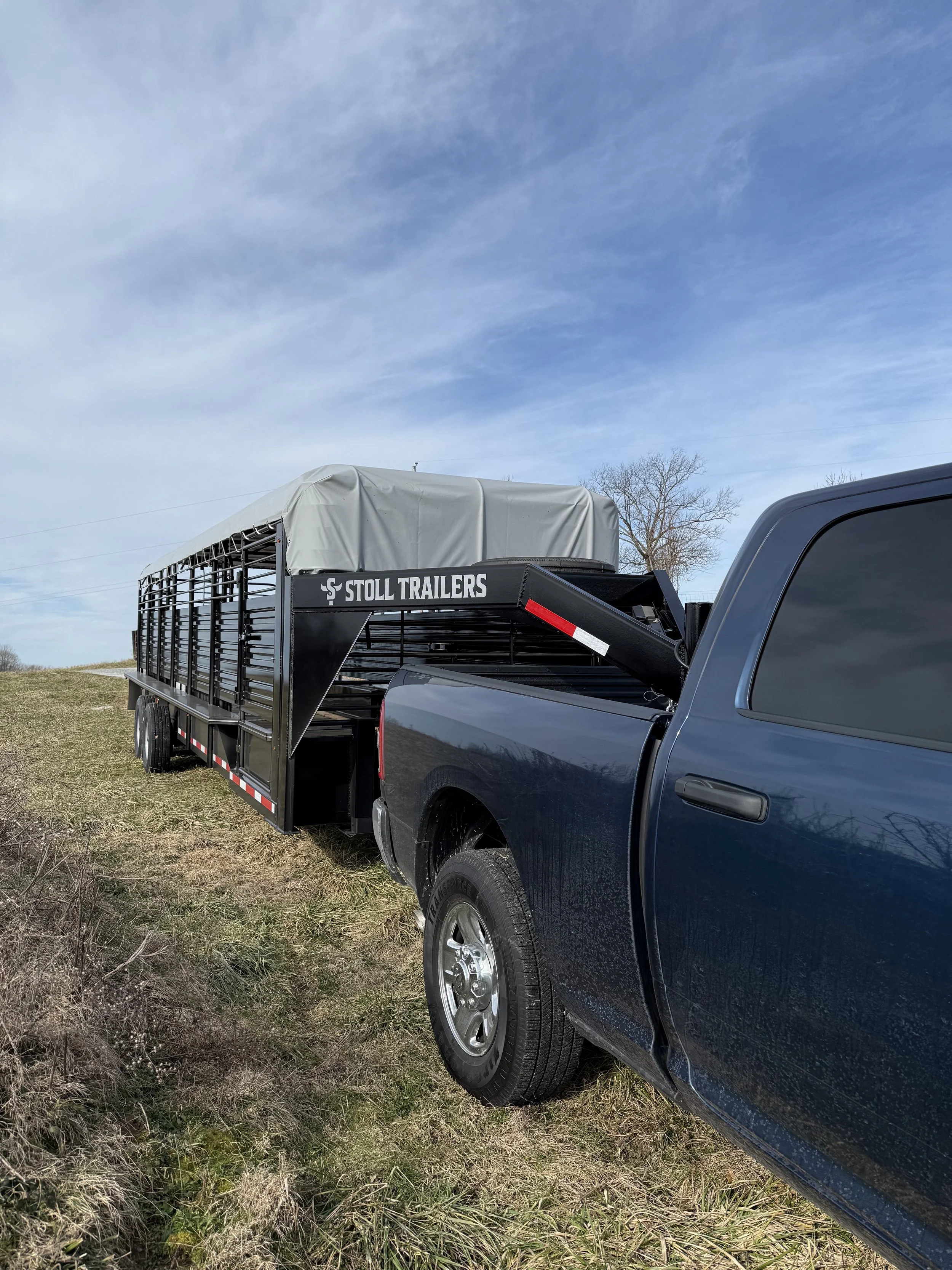 A black pickup truck with a trailer attached, parked on grass under a partly cloudy sky. The trailer has a canvas cover and is labeled 'Stoll Trailers'.